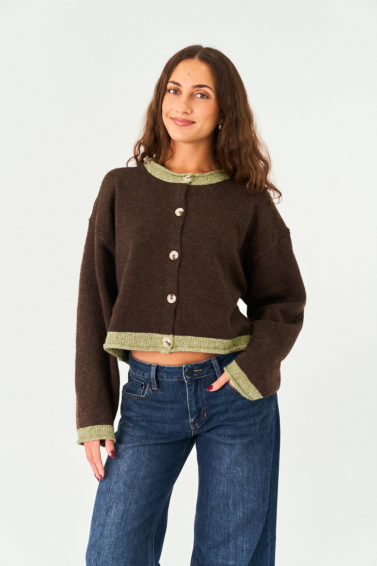 A woman modeling a cropped brown knit cardigan with sage green ribbed trim and silver buttons, paired with blue jeans, against a plain light gray studio background.