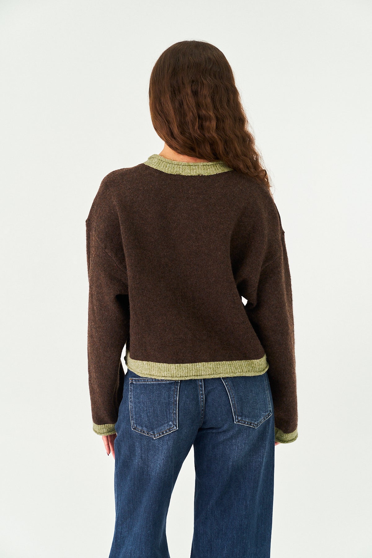 A woman viewed from behind wearing a dark brown knit sweater with light olive green ribbed trim at the crew neck, hem, and cuffs, paired with blue jeans against a plain white studio background.