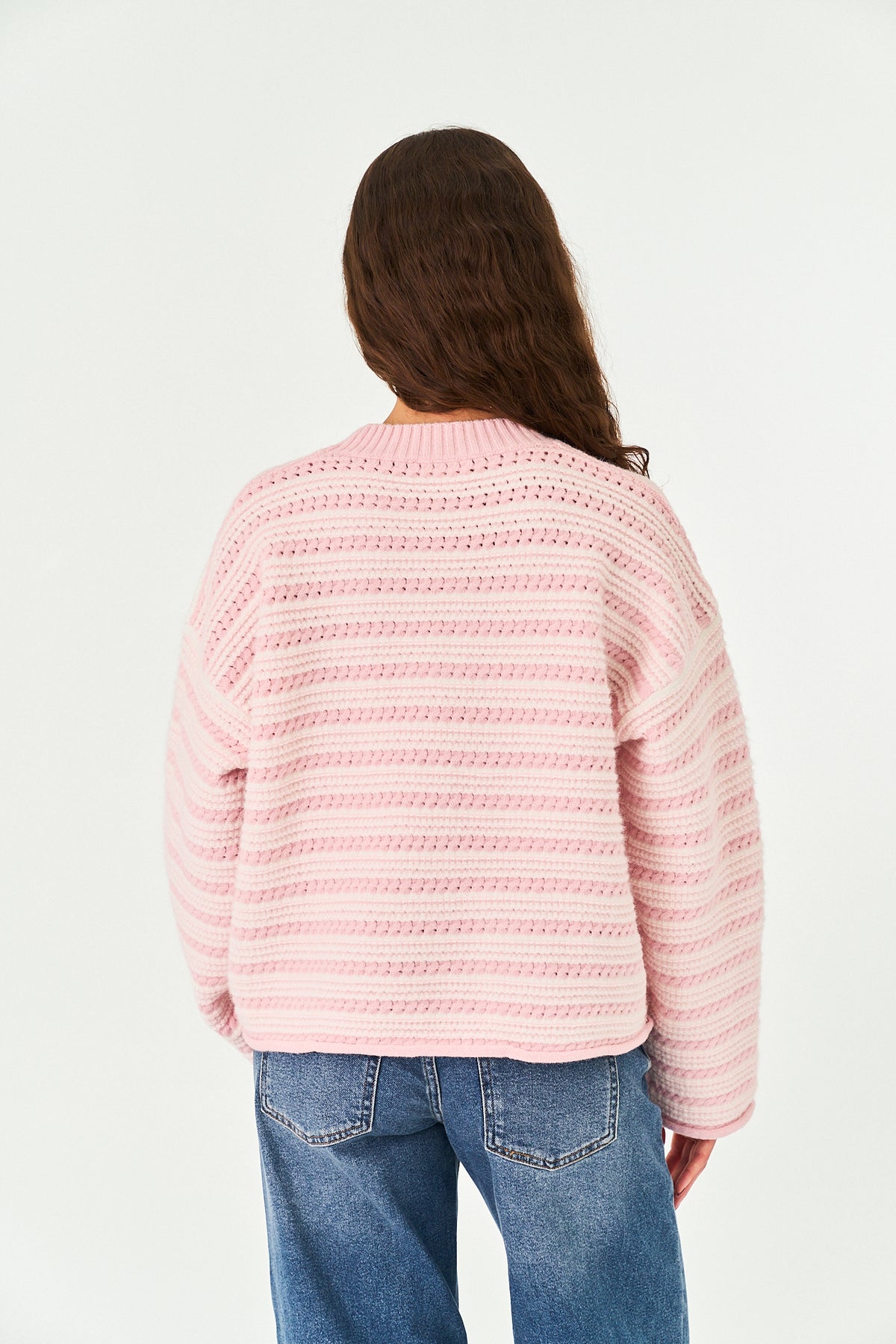A woman viewed from behind wearing a pink knit sweater with horizontal textured stripes and blue jeans, set against a plain white studio background.