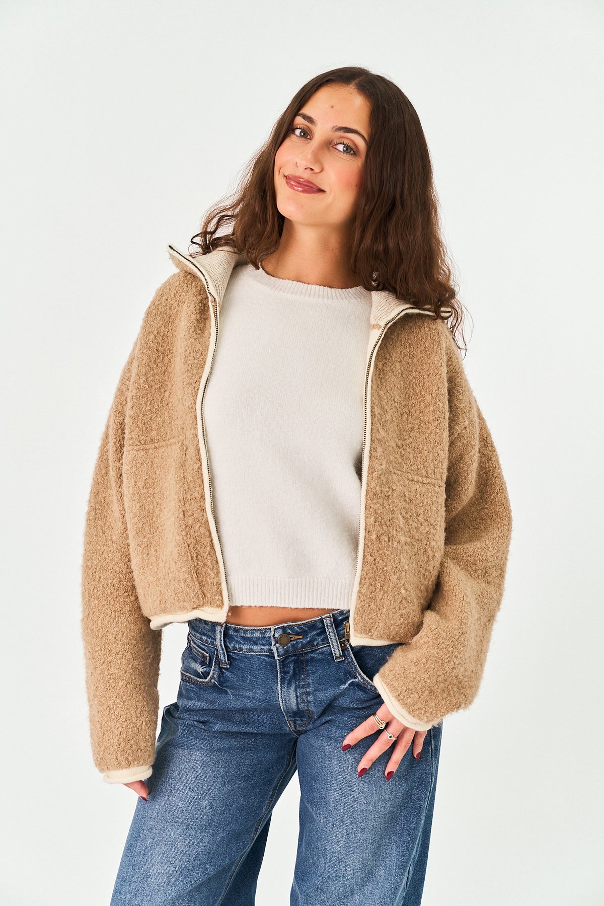 A woman wearing a cropped, high-collar teddy fleece jacket in a warm camel beige color with a cream zipper and cuffs, paired with blue jeans, against a plain white studio background.