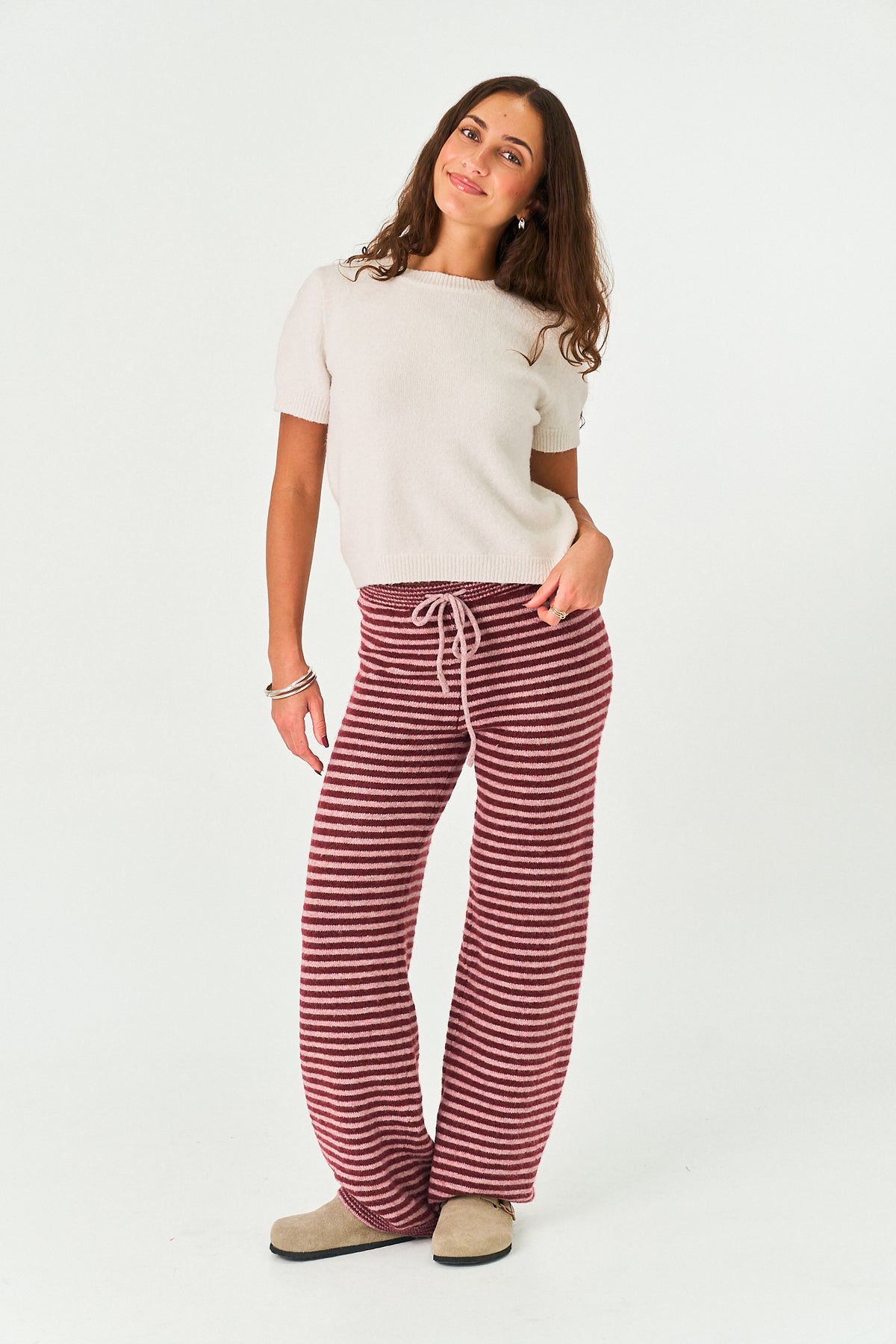 A woman modeling a pair of wide-leg knit pants with horizontal burgundy and white stripes, paired with a cream t-shirt and beige slippers, against a plain white studio background.
