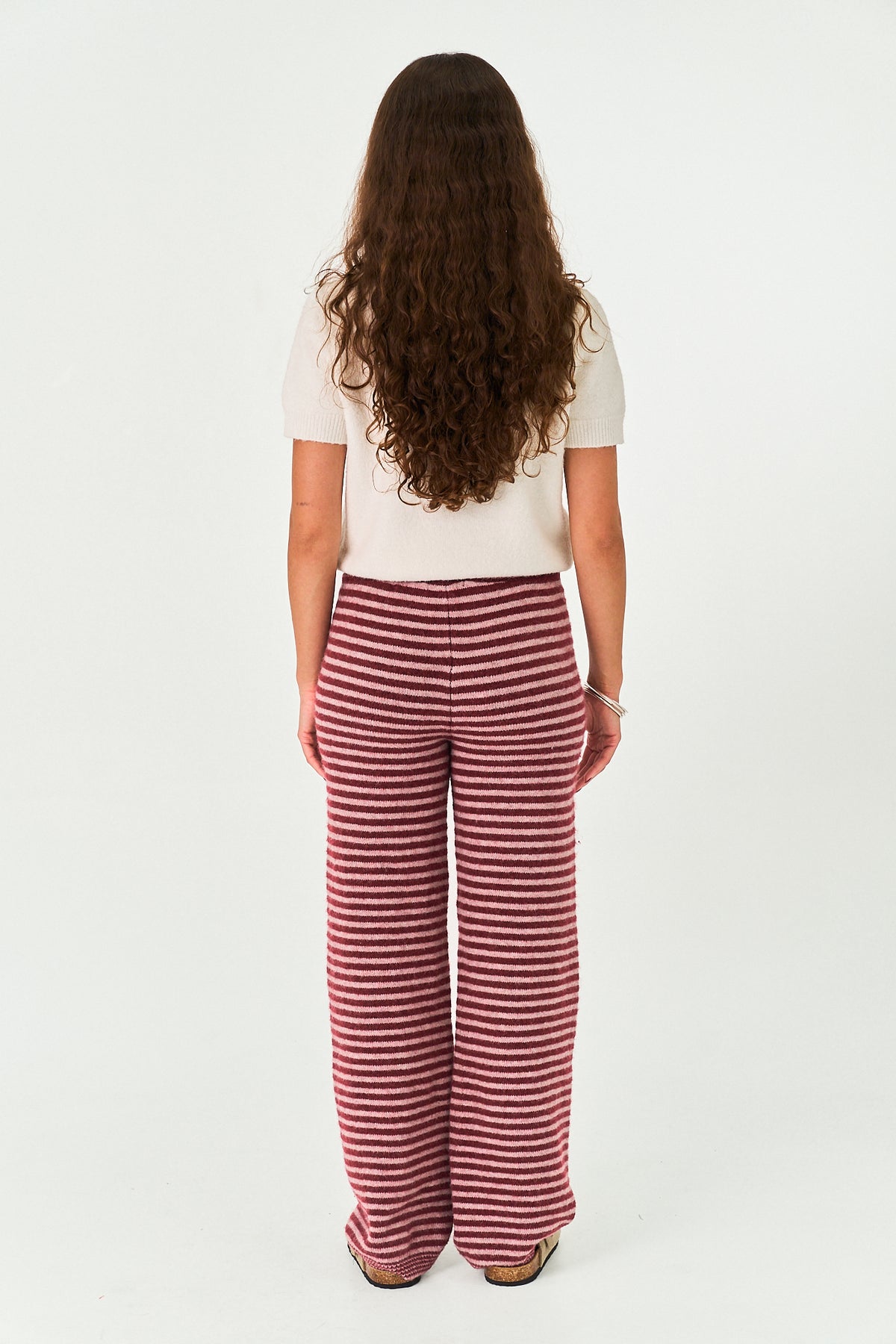 A woman viewed from behind wearing a white short-sleeve top and red-and-white horizontally striped wide-leg pants, standing against a plain white studio background.