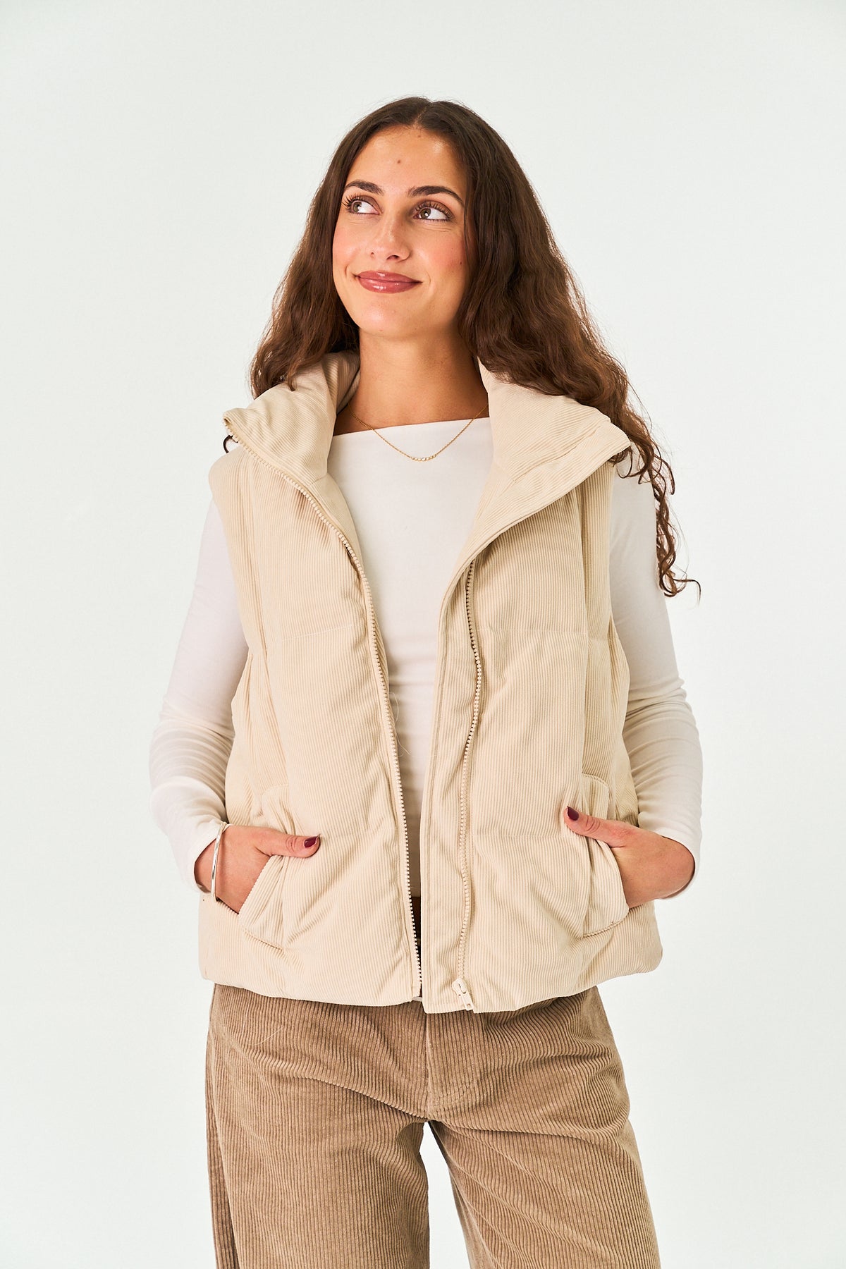 A woman wearing a light beige hooded puffer vest over a white long-sleeve top and brown corduroy pants, standing against a plain white studio background.