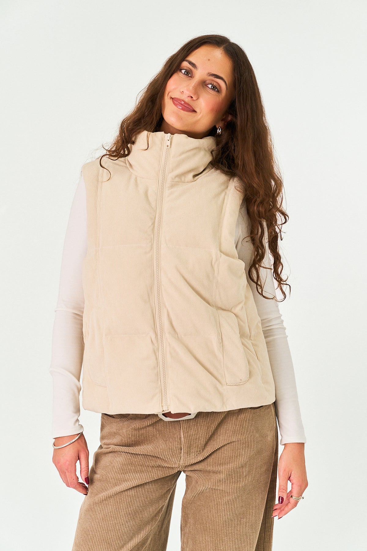 A woman wearing a sleeveless beige puffer vest over a white long-sleeve top and brown corduroy pants, photographed against a plain white studio background.