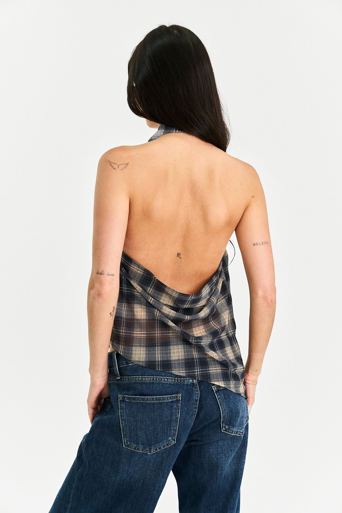 A woman shown from the back wearing a plaid halter top with an open back and dark blue jeans, against a plain light gray studio background.