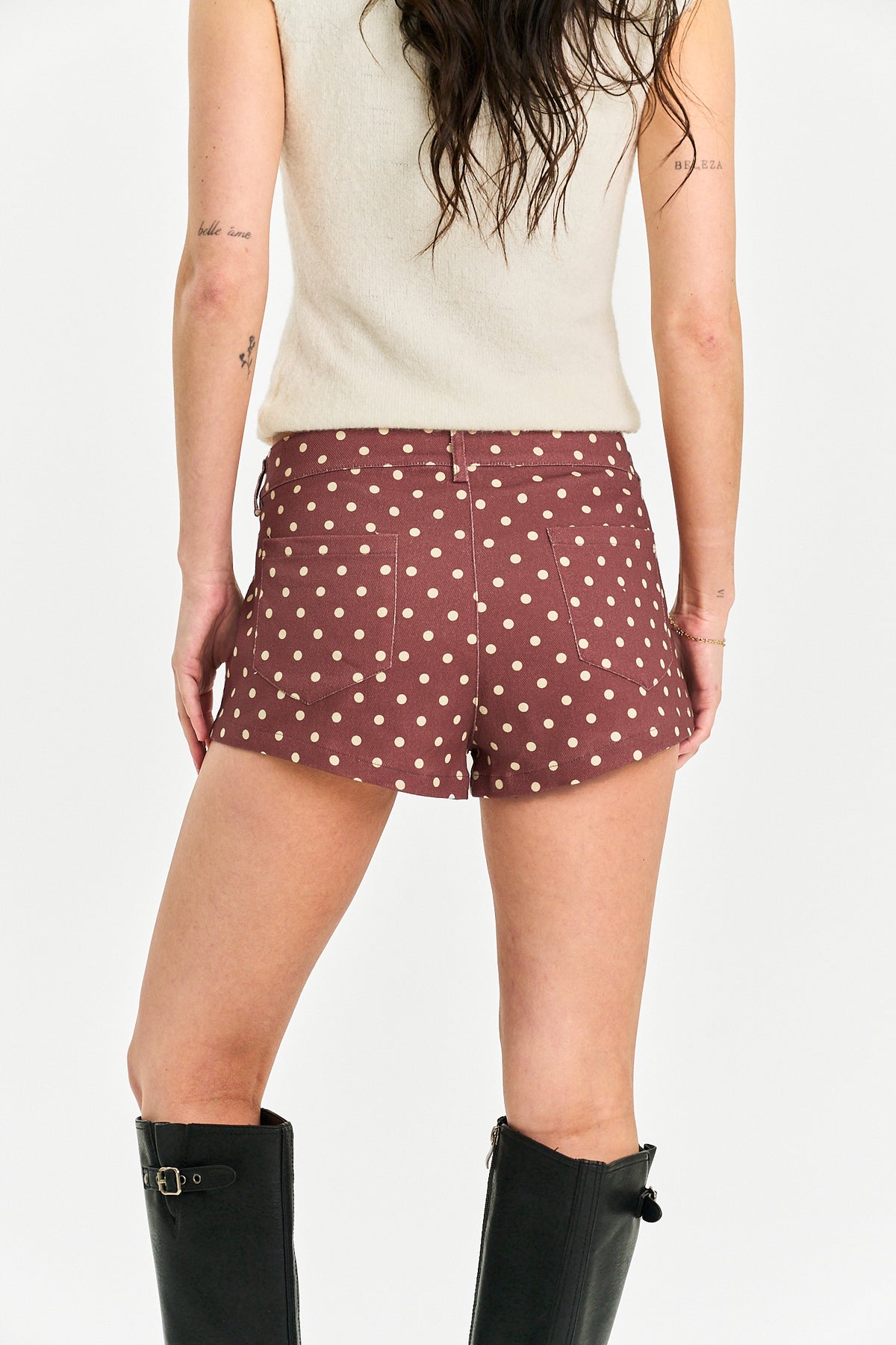 Rear view of a model wearing dusty rose shorts with a white polka dot pattern, paired with a cream sleeveless top and black knee-high boots against a light gray background.
