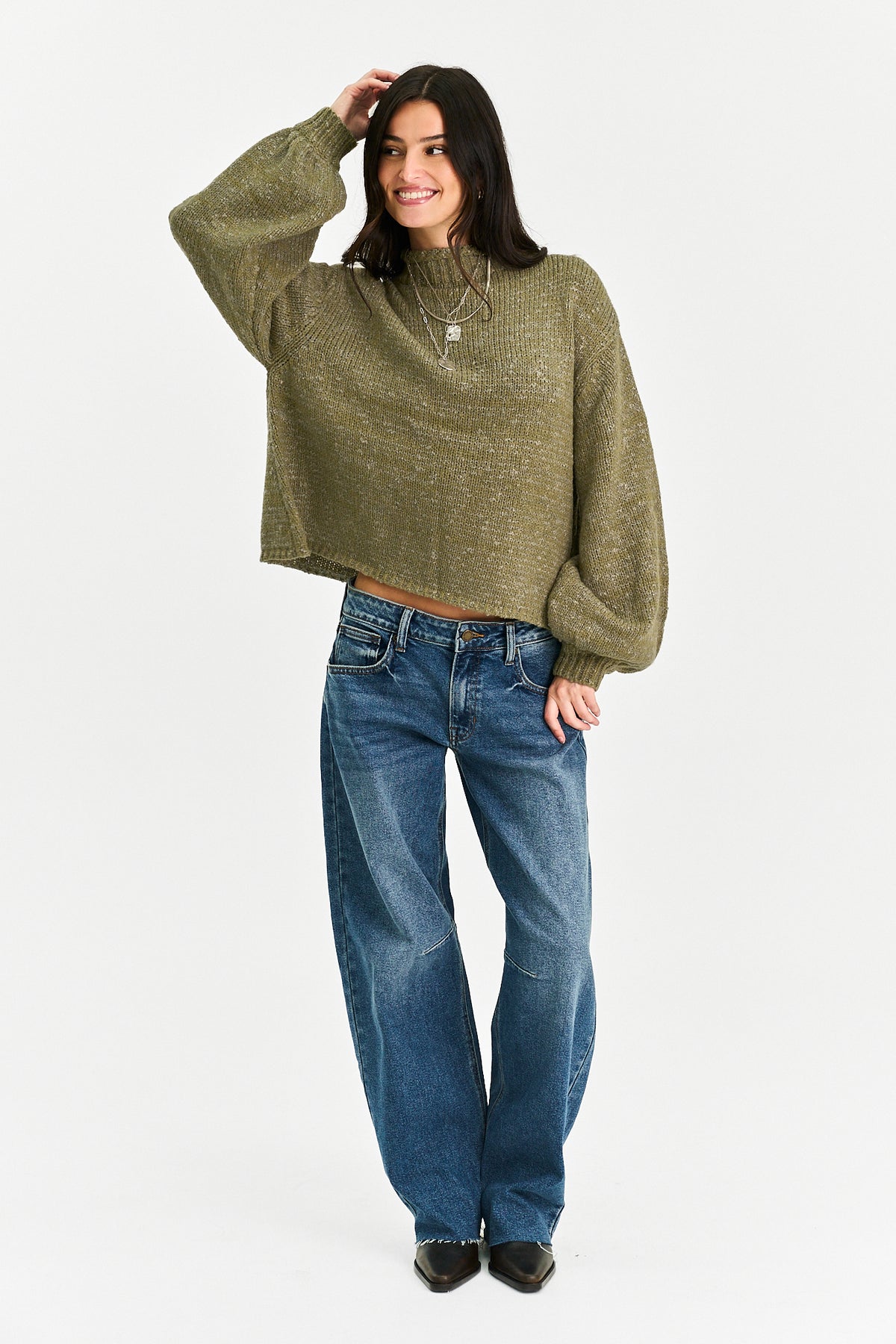 A woman wearing a textured olive green mock-neck knit sweater with balloon sleeves and blue jeans, standing against a light gray studio background.