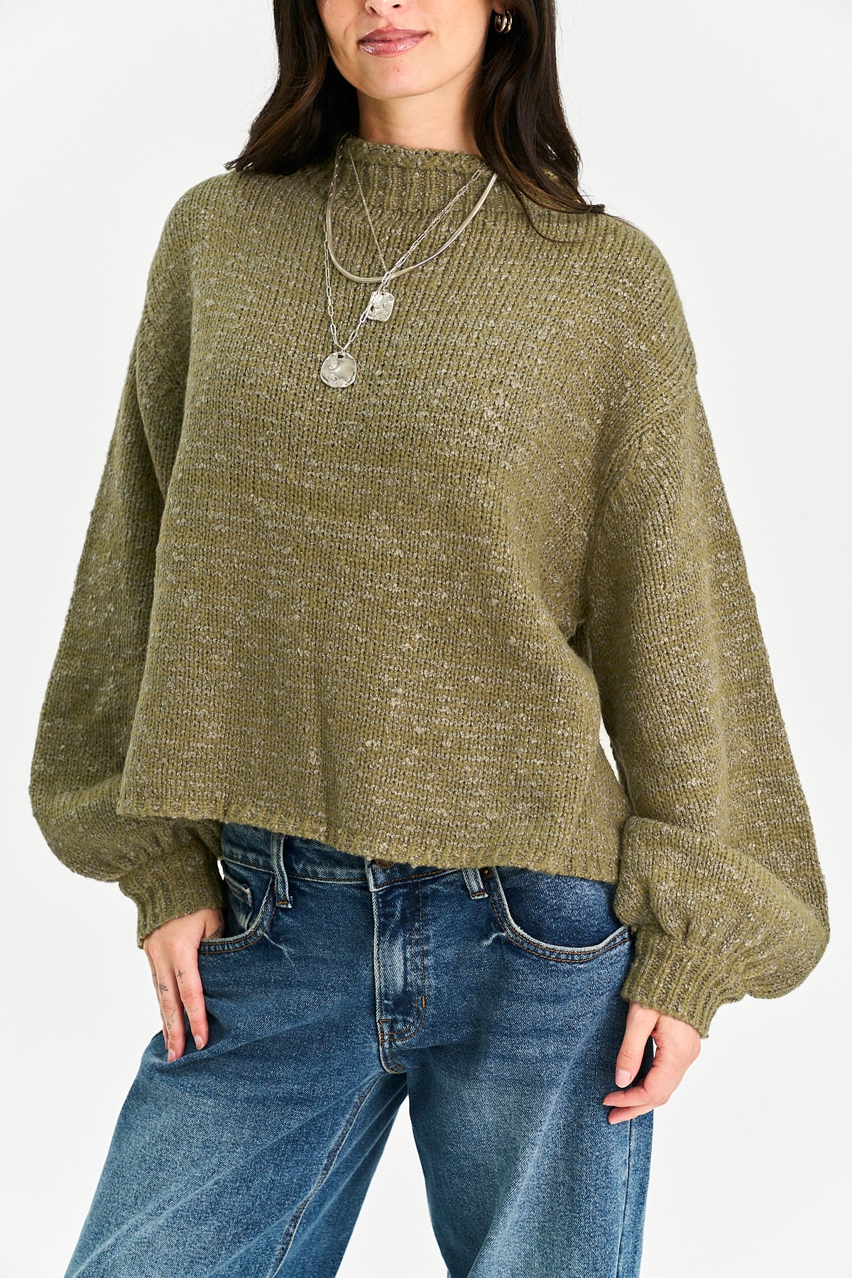 A woman wearing a textured olive green knit sweater with balloon sleeves and blue jeans, photographed against a clean white studio background.