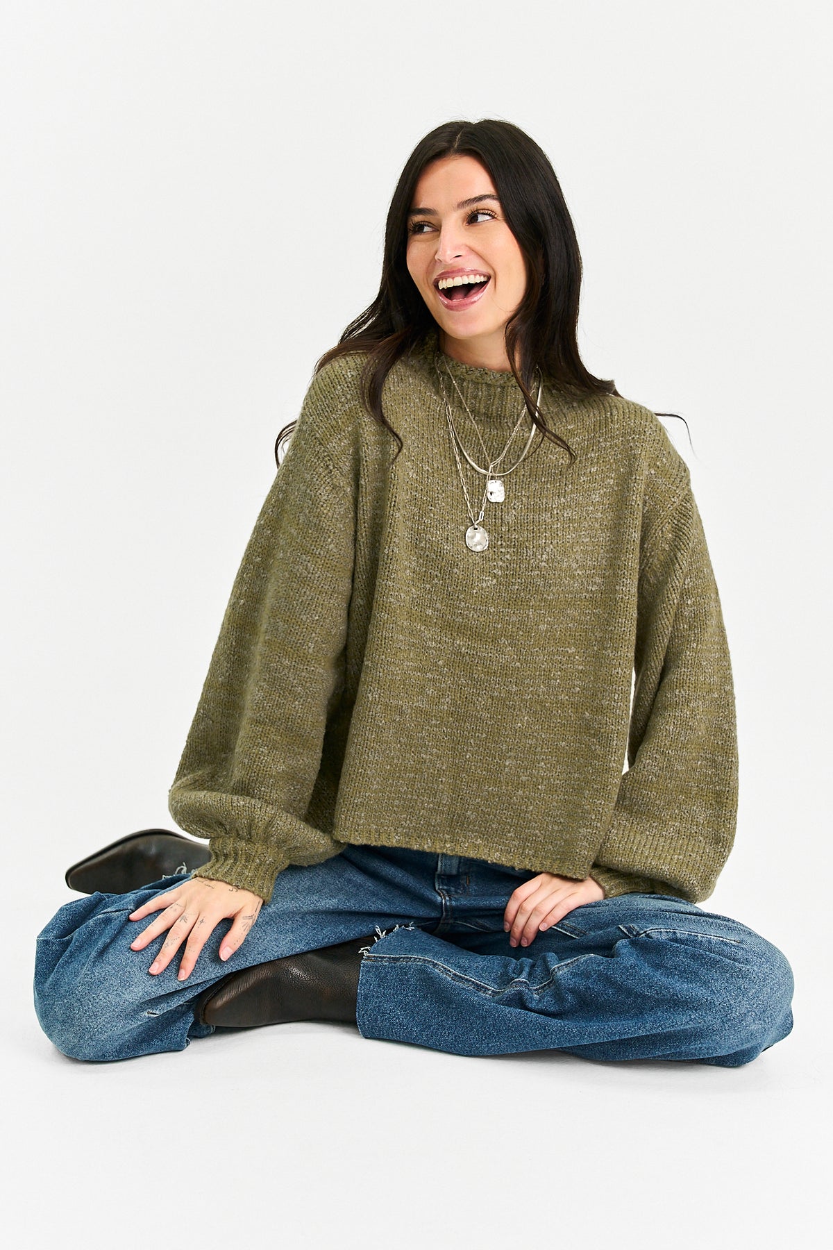 A woman sitting cross-legged on a light gray studio background, wearing a chunky olive green knit sweater, blue jeans, and black boots.