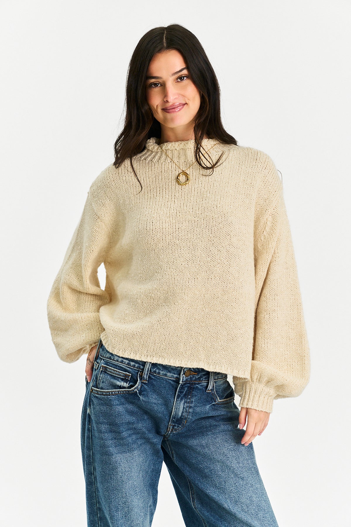 A woman wearing a cream-colored, textured knit sweater with balloon sleeves and a gold circular pendant at the collar, paired with blue jeans, against a plain light gray background.