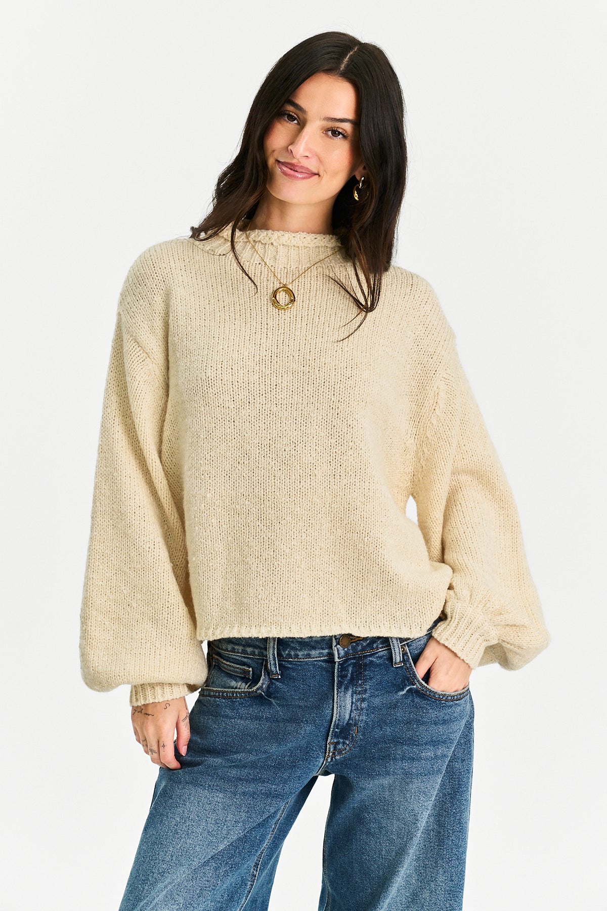 A woman wearing a cream-colored, textured knit sweater with balloon sleeves and blue jeans, standing against a plain white background.