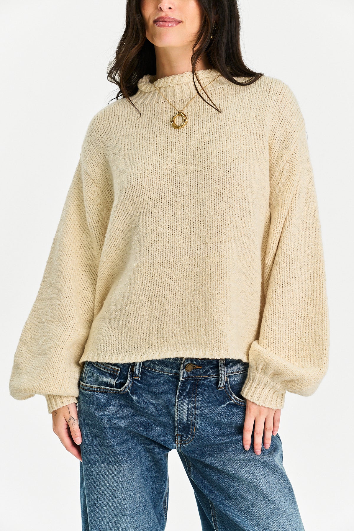 A woman wearing a cream-colored, chunky knit mock-neck sweater with balloon sleeves, paired with blue jeans, against a plain white background.