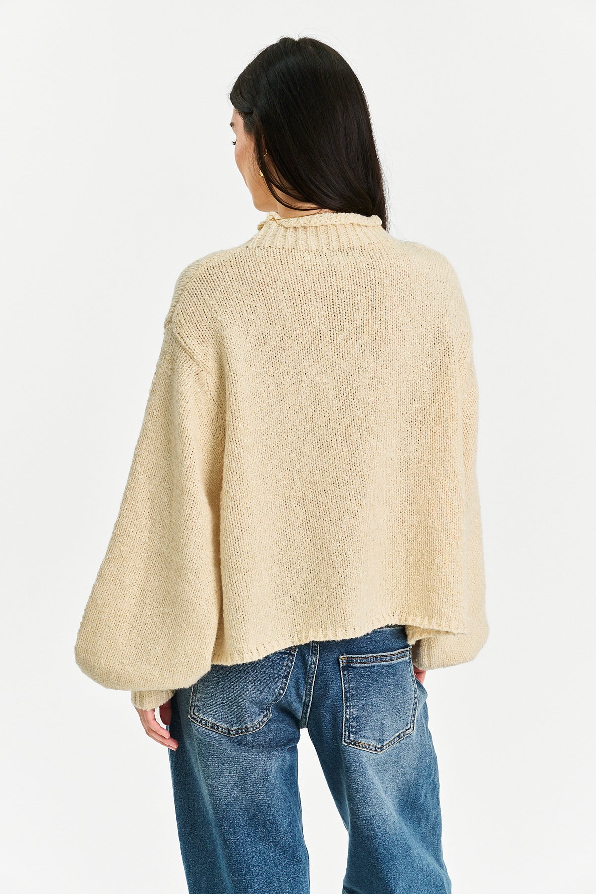 A woman shown from the back wearing a cream-colored, textured knit mock-neck sweater with wide balloon sleeves, paired with blue jeans, against a plain white background.