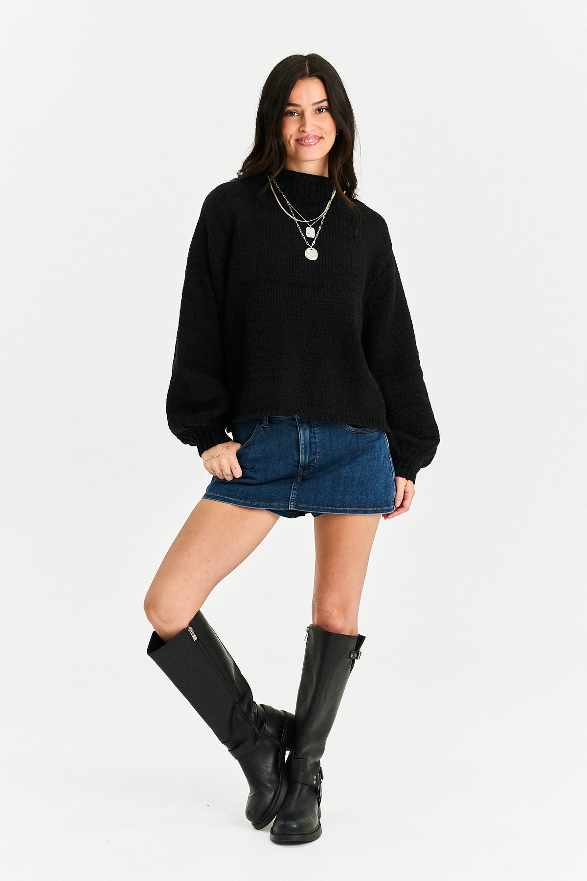 A woman modeling a black knit mock-neck sweater, paired with a denim mini skirt, layered silver necklaces, and black knee-high leather boots, against a plain white studio background.