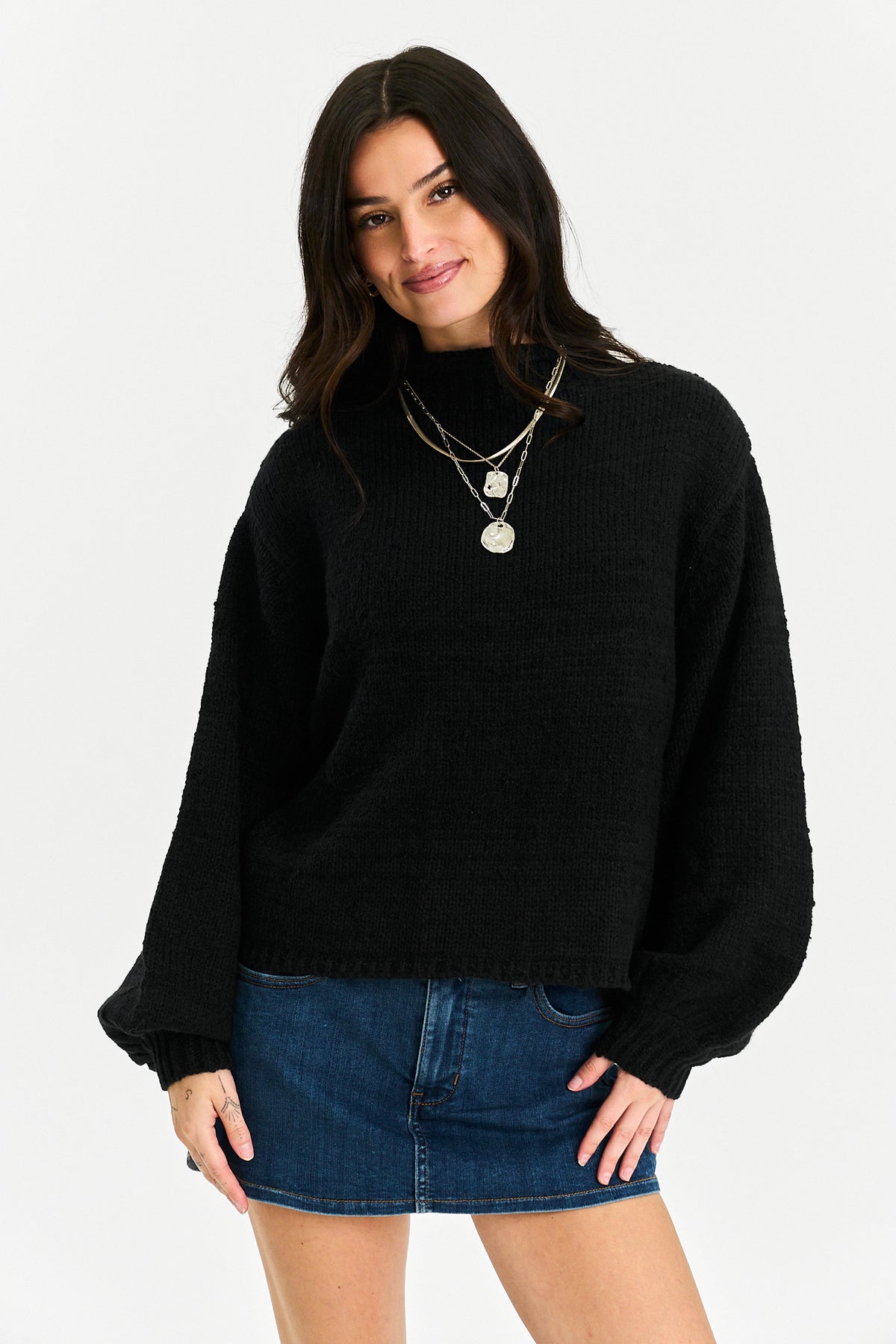 A woman modeling a black ribbed knit mock-neck sweater with balloon sleeves, paired with a blue denim mini skirt and layered silver necklaces, against a plain white studio background.
