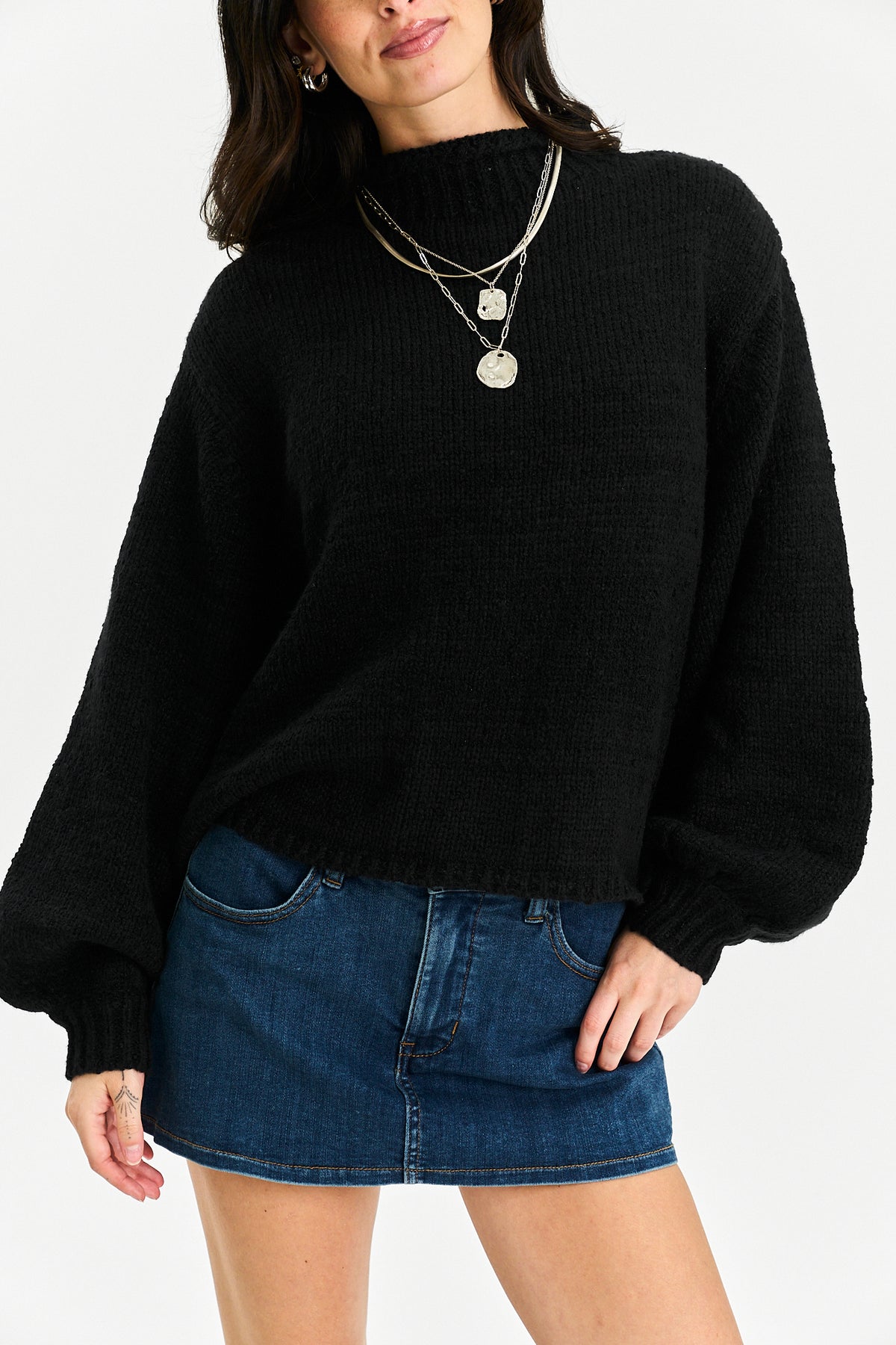 A woman wearing a black ribbed knit mock-neck sweater with balloon sleeves and a dark blue denim mini skirt, accessorized with layered silver necklaces, against a plain white background.