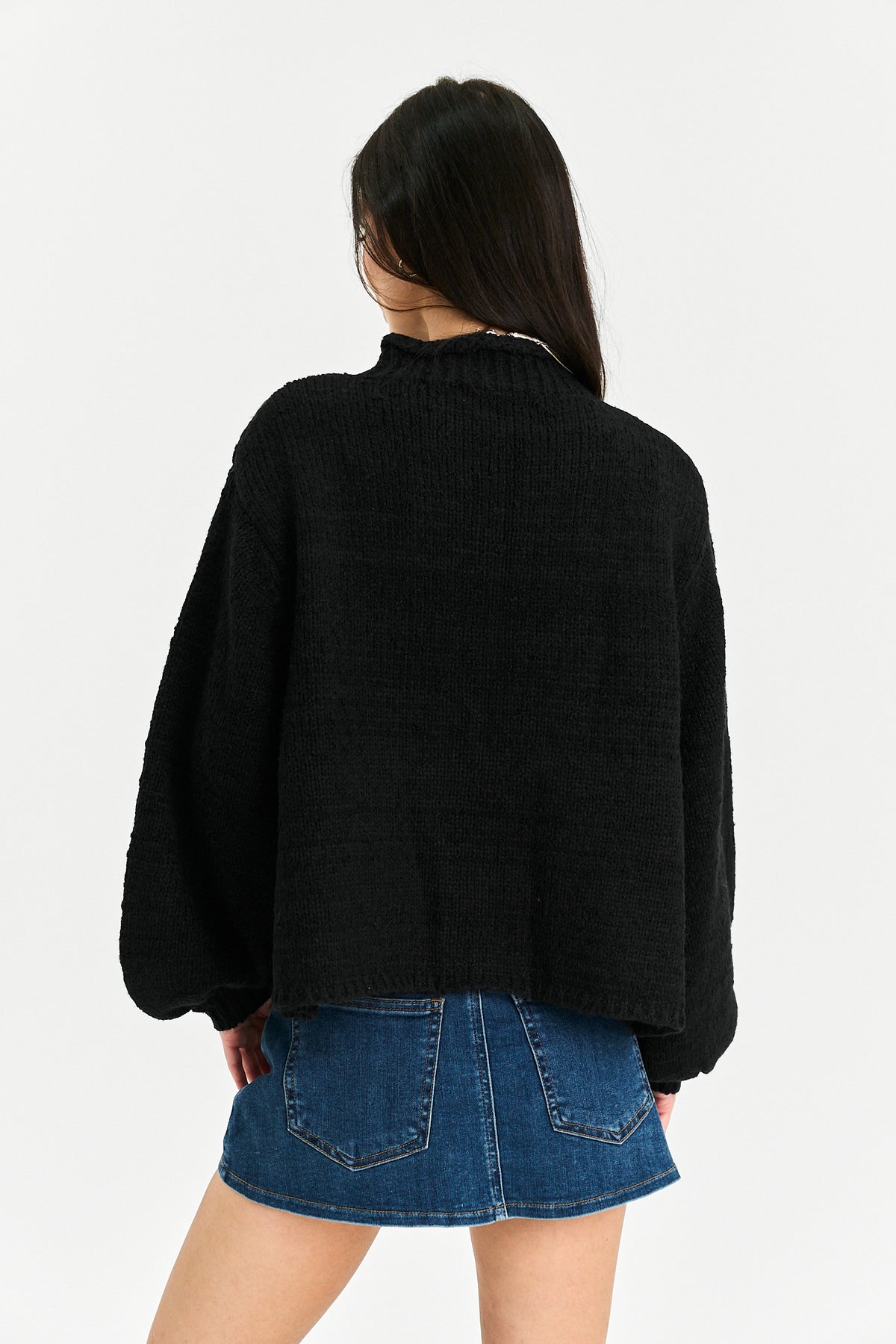 A woman shown from the back wearing a black cropped knit sweater with balloon sleeves and a high ribbed collar, paired with a blue denim skirt, against a plain light gray studio background.