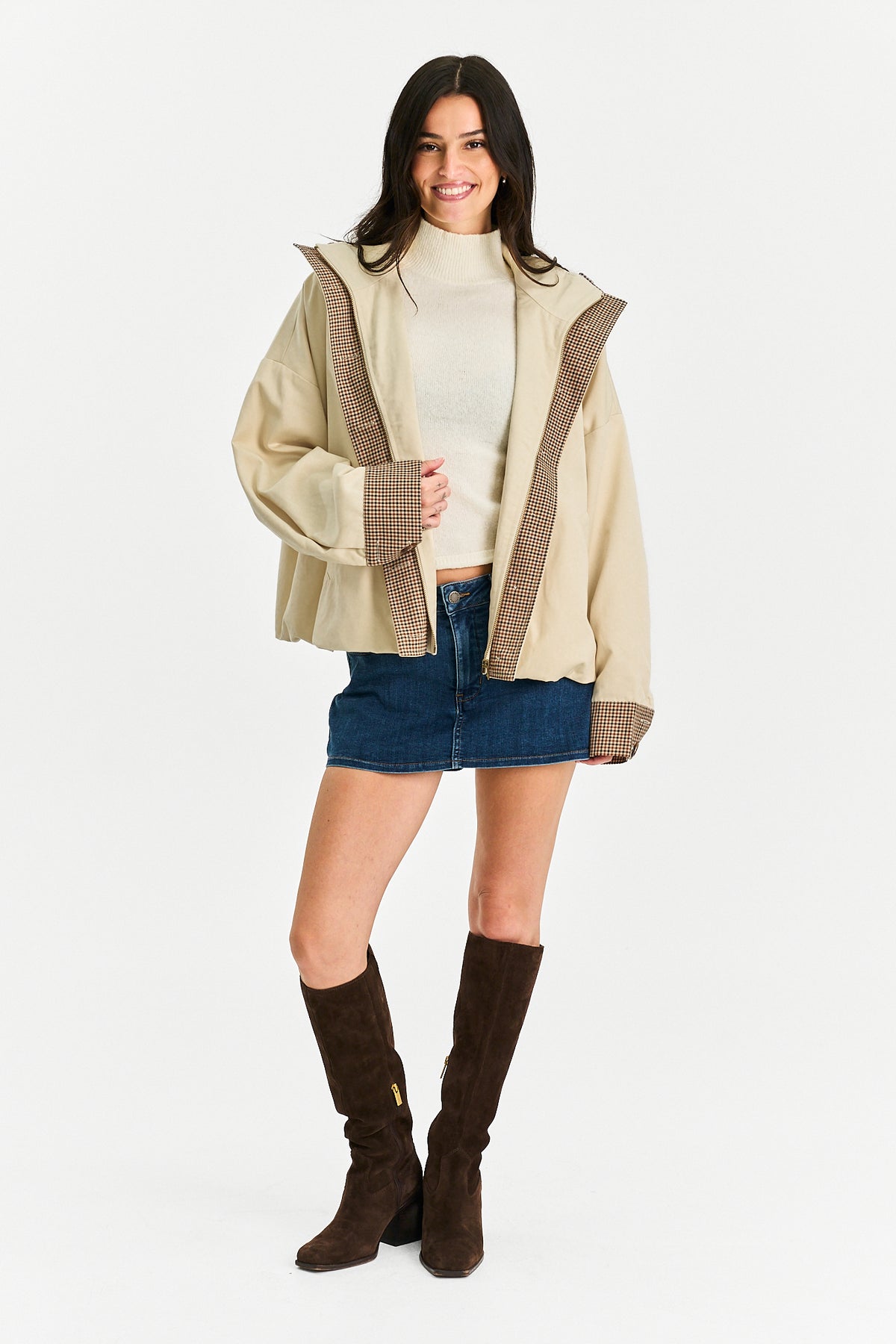 A woman modeling a beige oversized jacket with brown plaid trim, worn over a cream turtleneck, blue denim mini skirt, and dark brown knee-high boots, against a plain white studio background.