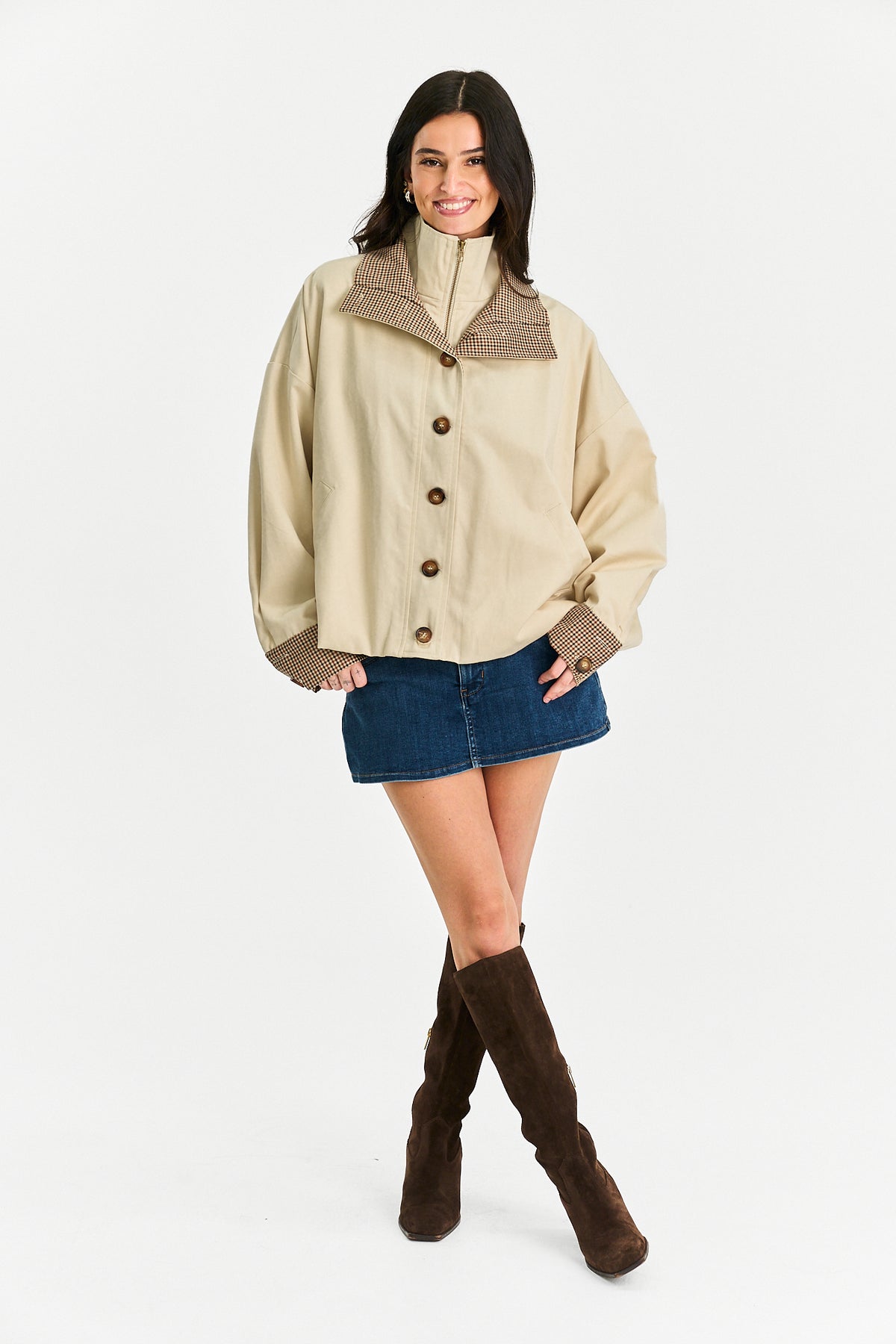 A woman modeling a beige cropped jacket with brown textured trim and buttons, paired with a blue denim mini skirt and brown knee-high boots, against a plain white studio background.