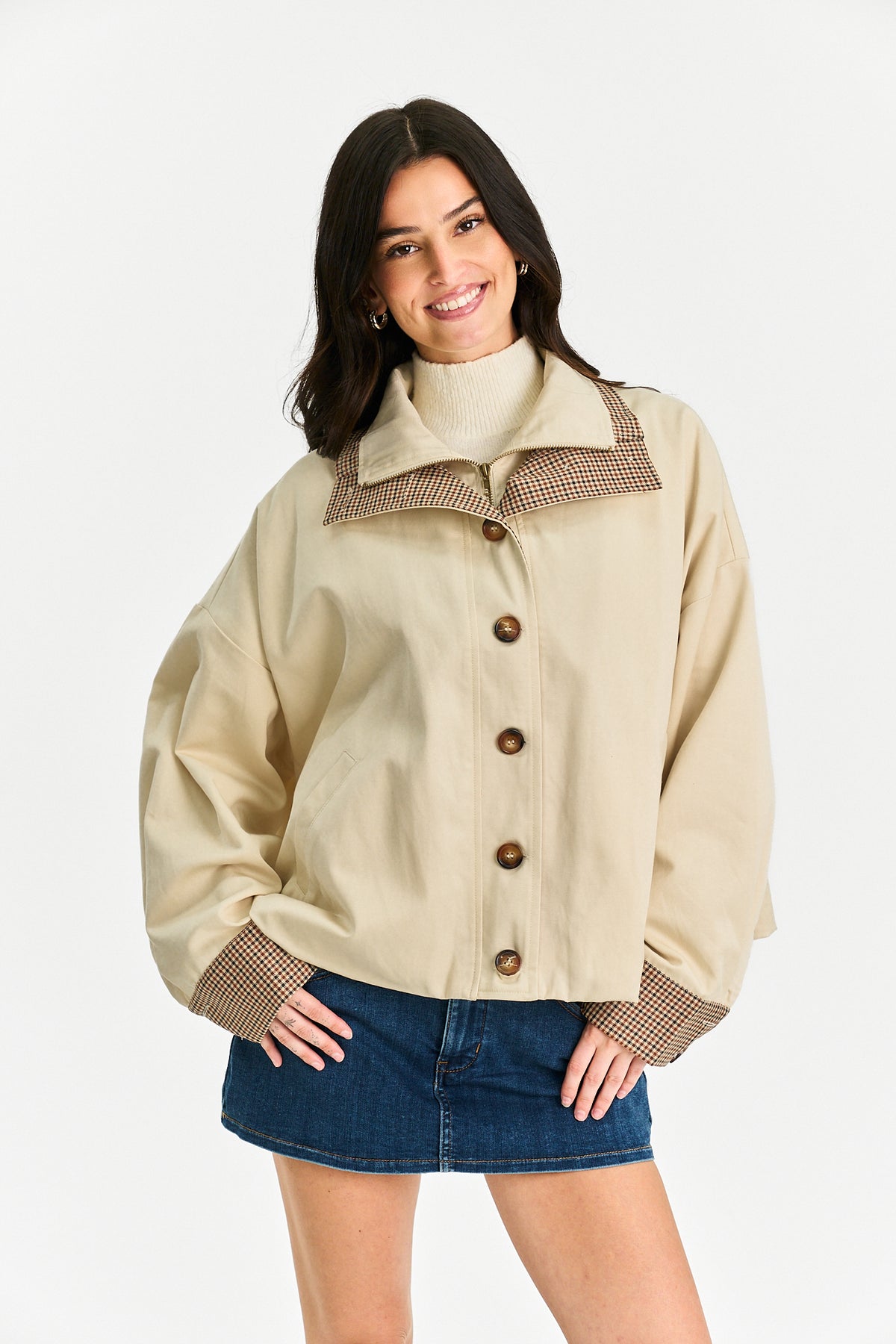 A woman modeling a beige utility-style jacket with brown and beige patterned trim on the collar and cuffs, paired with a blue denim skirt, against a plain white studio background.