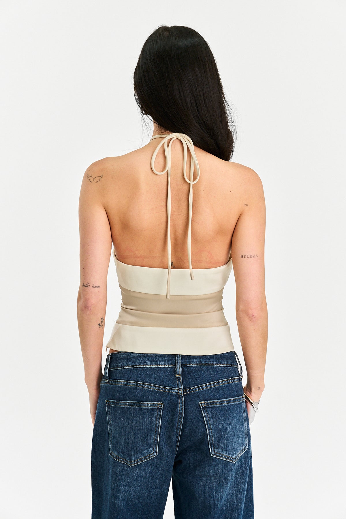 A woman shown from the back wearing a beige halter-style wrap top with a tie at the back and blue jeans, against a plain light gray studio background.