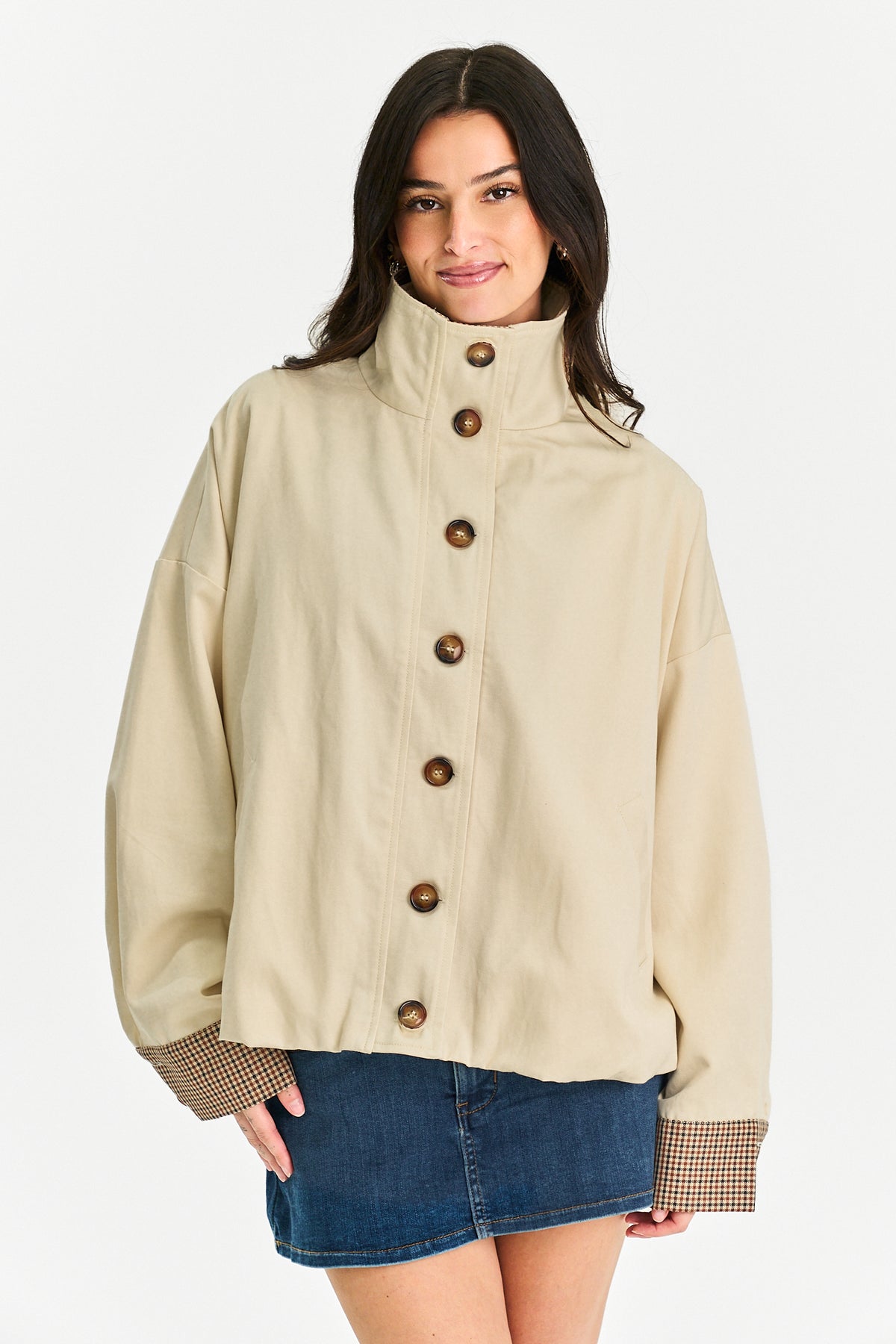 A woman modeling a beige high-neck utility jacket with large brown buttons and patterned cuffs, paired with a denim mini skirt, against a plain white studio background.