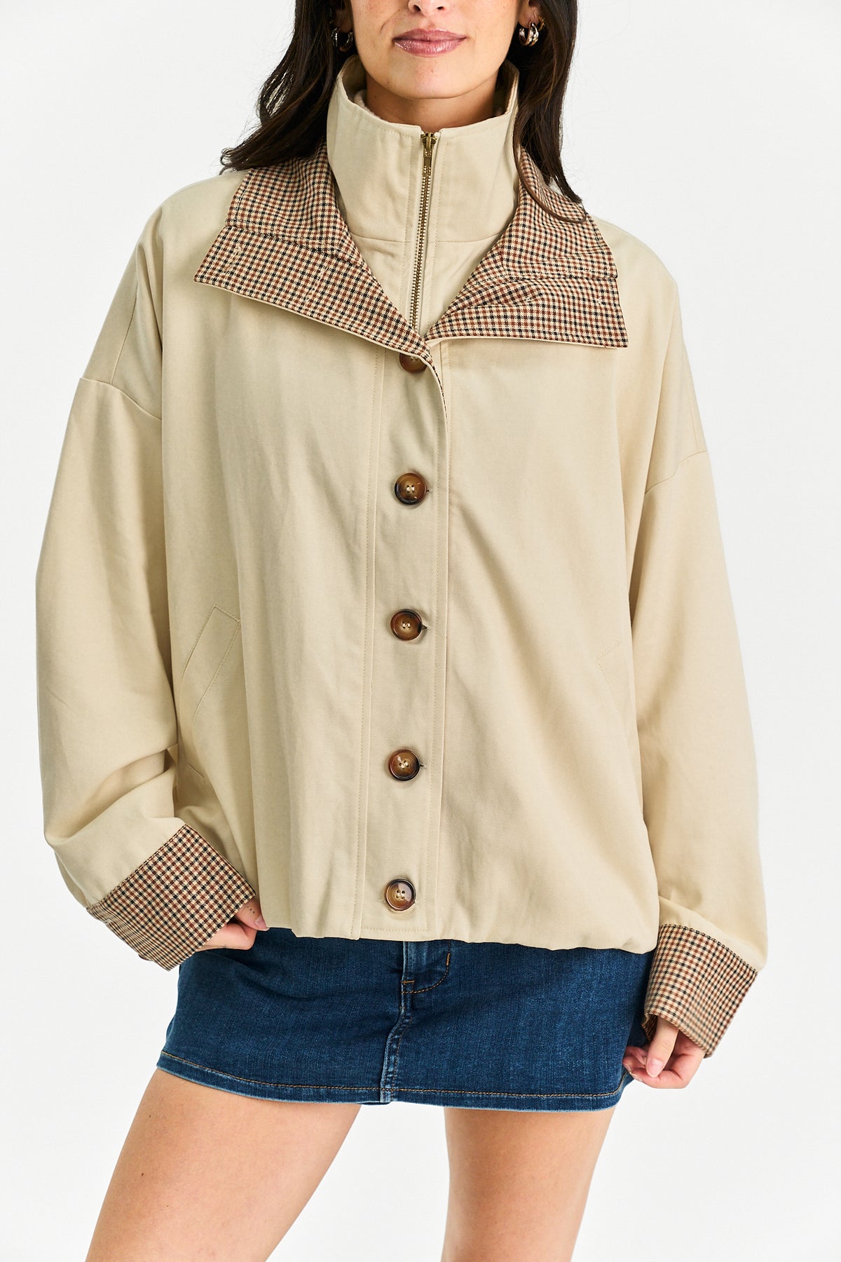 A woman wearing a beige cropped jacket with a high collar and brown-and-beige checkered trim, paired with a blue denim skirt, against a plain light background.