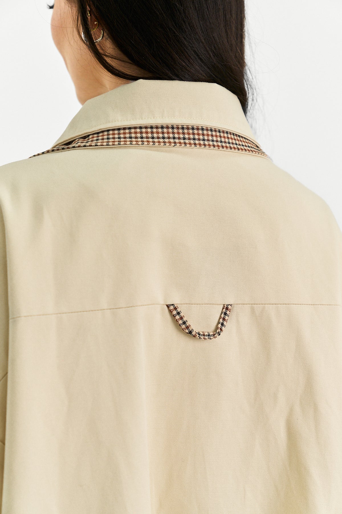 Close-up back view of a person wearing a light beige shirt jacket with a plaid patterned collar and matching plaid detail near the lower back, against a plain light background.