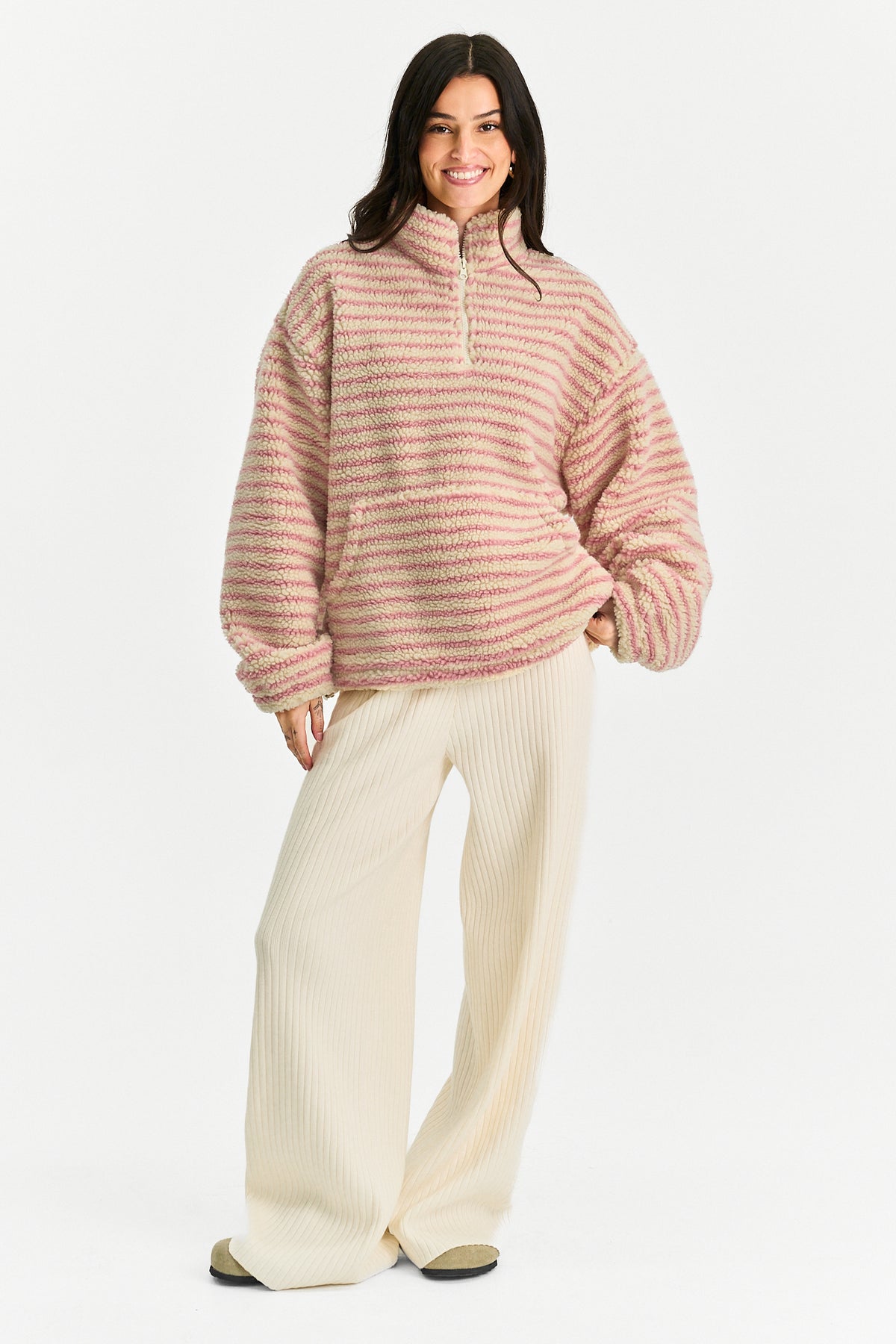 A woman modeling a fuzzy quarter-zip pullover sweater with horizontal pink and cream stripes, paired with cream wide-leg knit pants, against a plain white studio background.