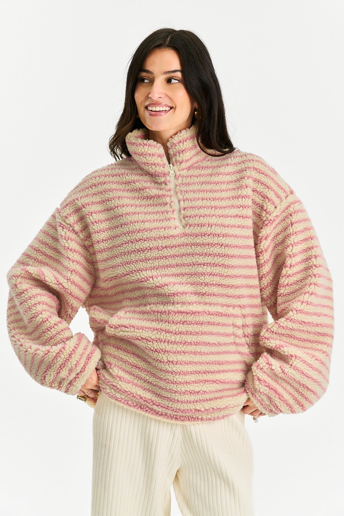A woman wearing a fuzzy quarter-zip pullover sweater with horizontal pink and cream stripes, paired with cream ribbed pants, against a plain light gray studio background.