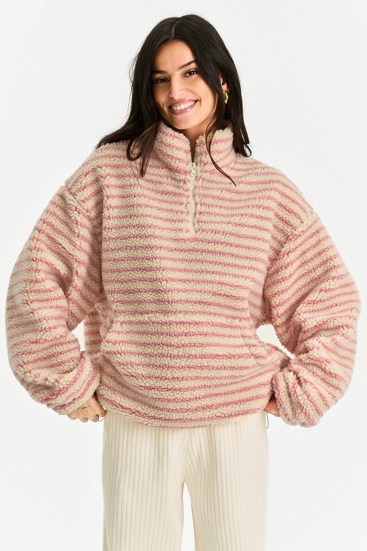 A woman wearing a fuzzy quarter-zip pullover sweater with horizontal pink and cream stripes, paired with cream ribbed pants, against a plain white background.
