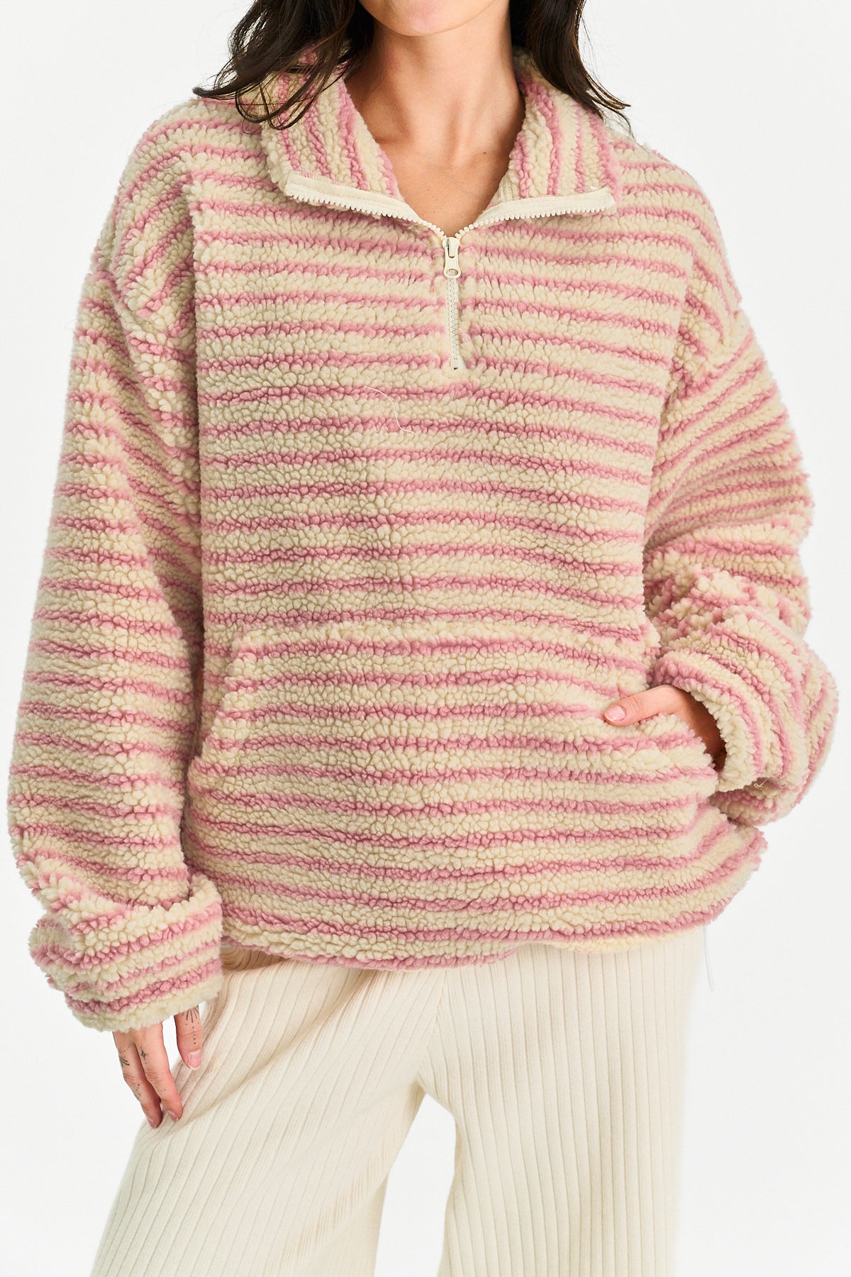 A person wearing a fuzzy quarter-zip pullover sweater with horizontal pink and cream stripes, paired with cream ribbed pants, against a plain light background.
