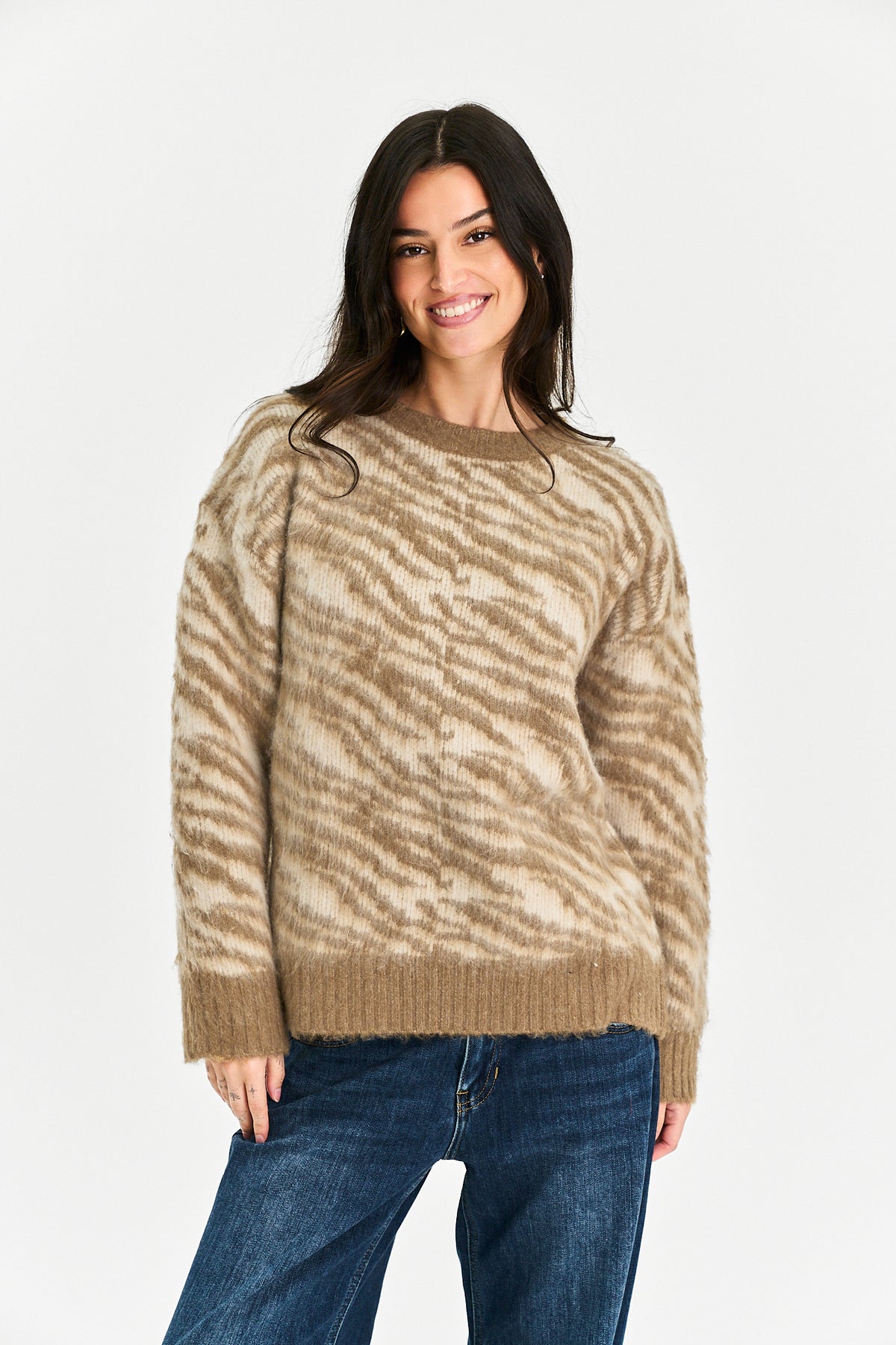 A woman modeling a beige and brown zebra-patterned knit sweater with blue jeans, photographed against a clean white studio background.