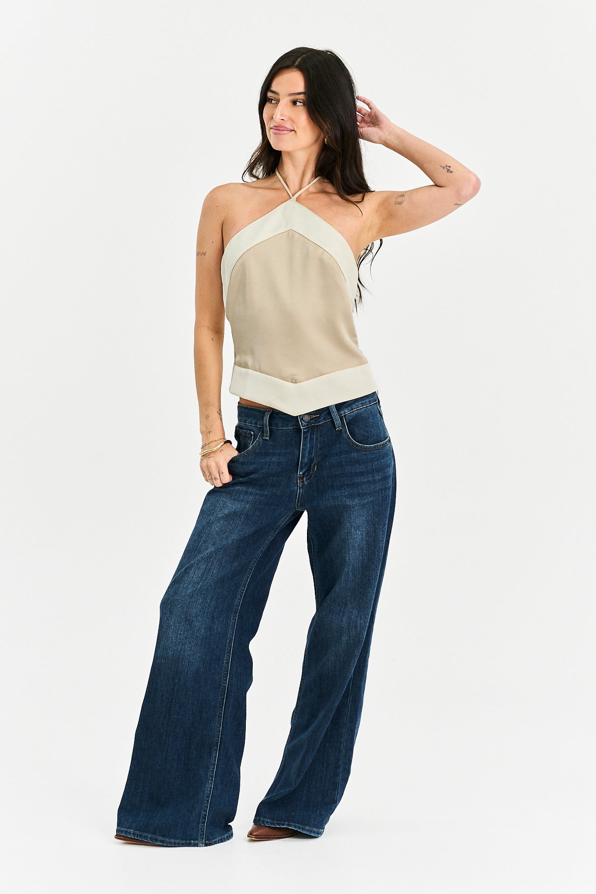 A woman modeling a beige and cream halter-neck knit top paired with dark blue wide-leg jeans, standing against a plain white studio background.