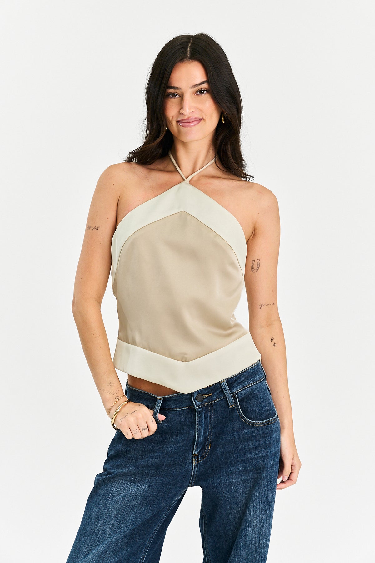 A woman modeling a beige halter crop top with contrasting cream panels and blue jeans against a plain white studio background.
