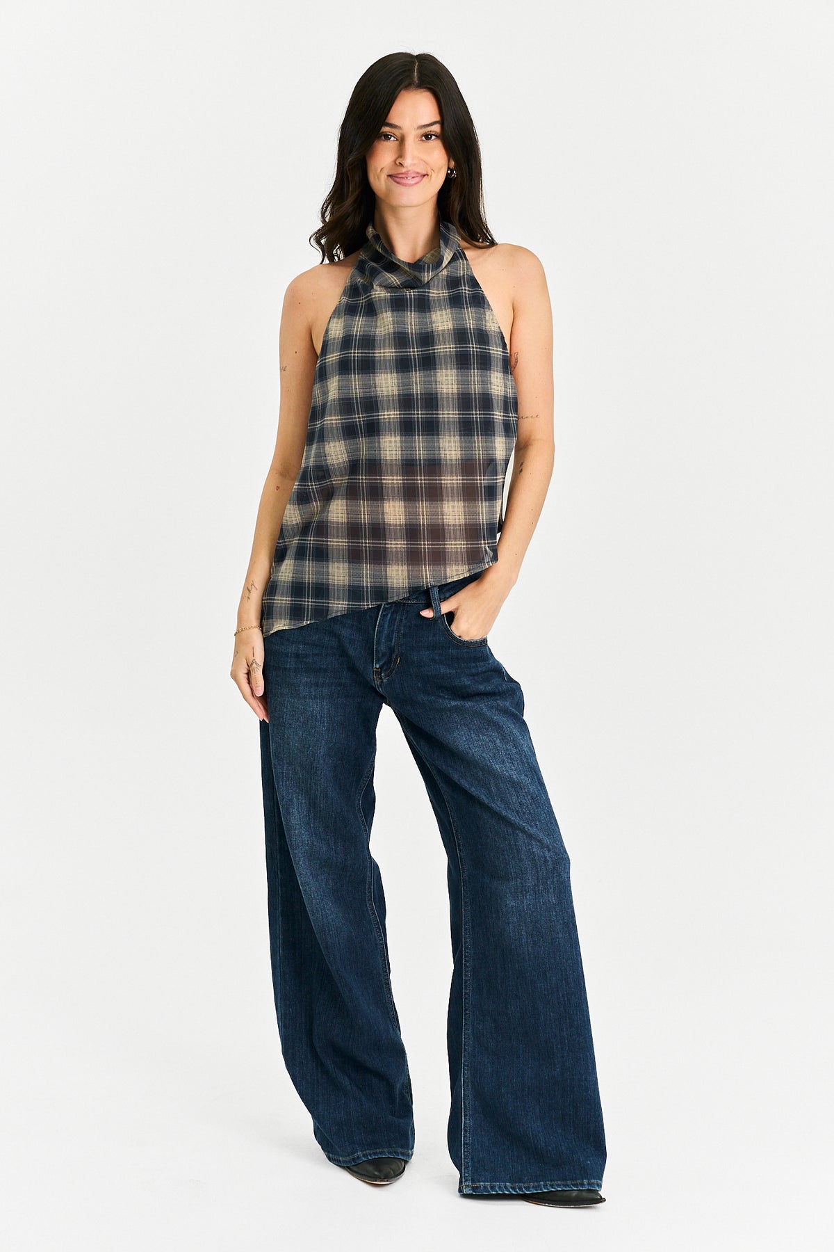 A woman modeling a sleeveless halter-neck plaid top in beige, brown, and navy, paired with dark blue wide-leg jeans, against a plain white studio background.