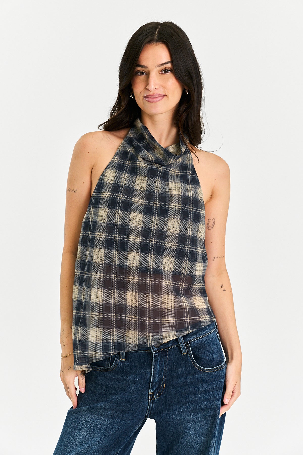 A woman modeling a sleeveless halter-neck top with a dark navy and beige plaid pattern, paired with blue jeans, against a plain light gray background.