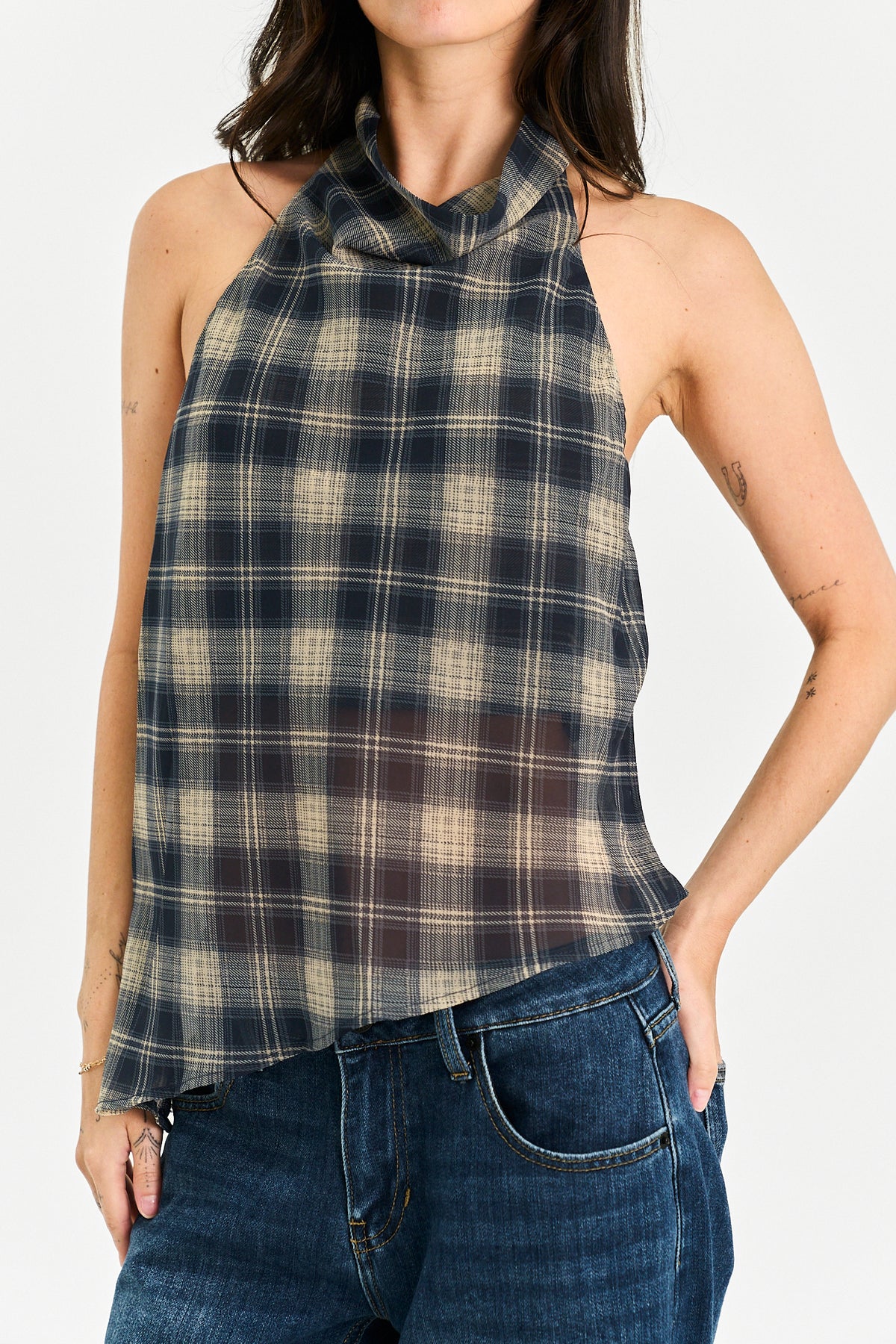 A woman wearing a sleeveless halter-neck plaid top in dark navy and beige, paired with blue jeans, against a plain light background.