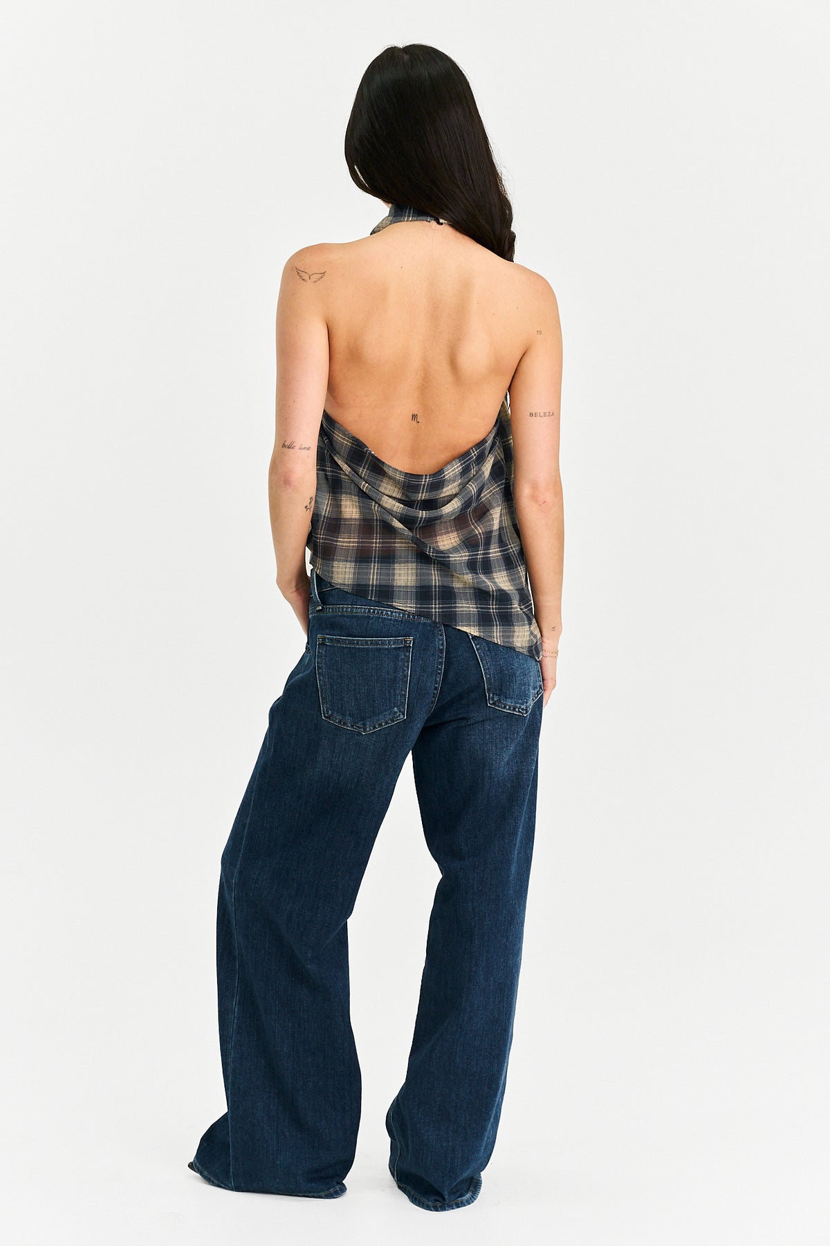 A woman shown from the back wearing a plaid halter-style top and dark wide-leg jeans against a plain light gray studio background.