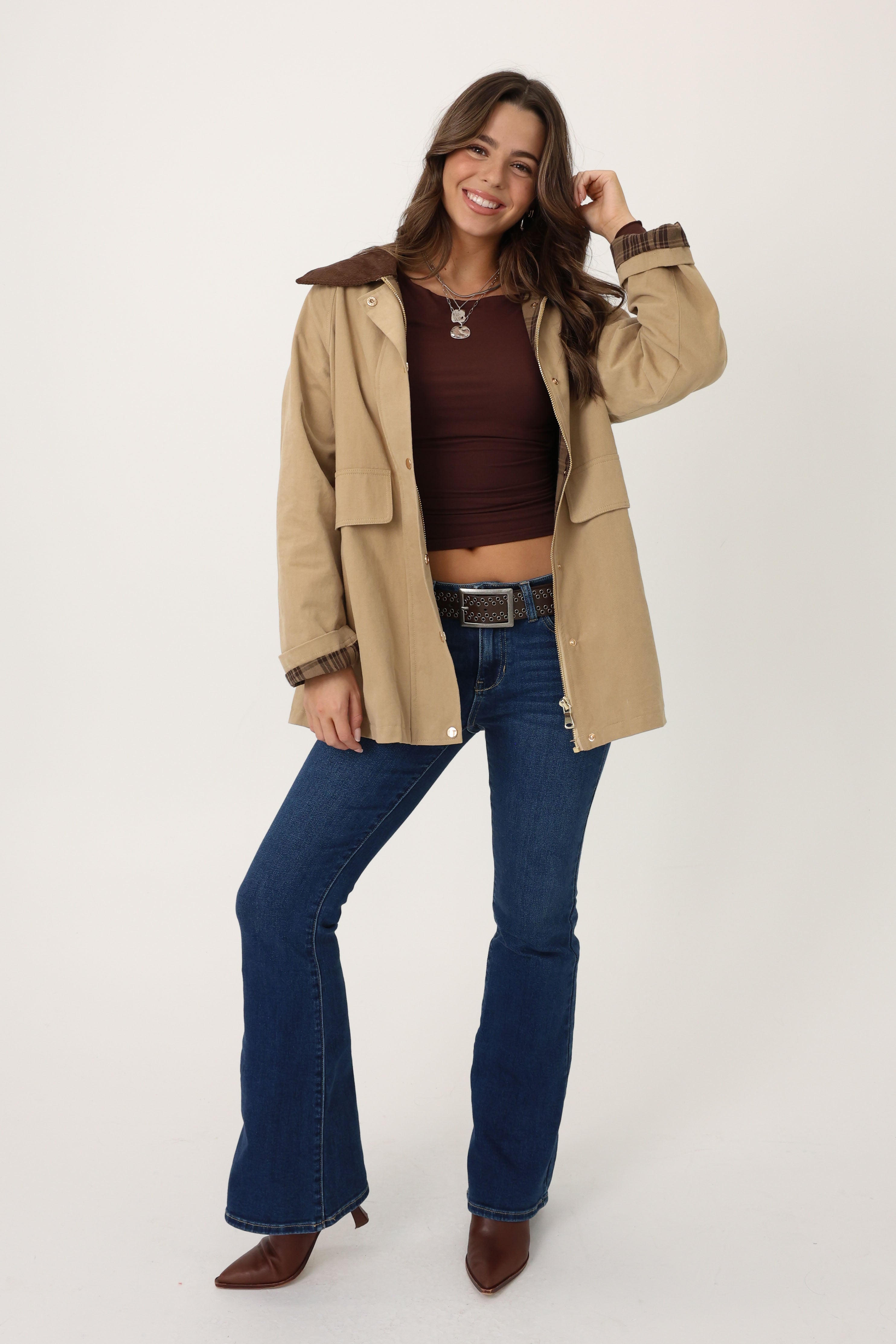 Woman wearing a beige jacket, brown top, and blue jeans on a white background