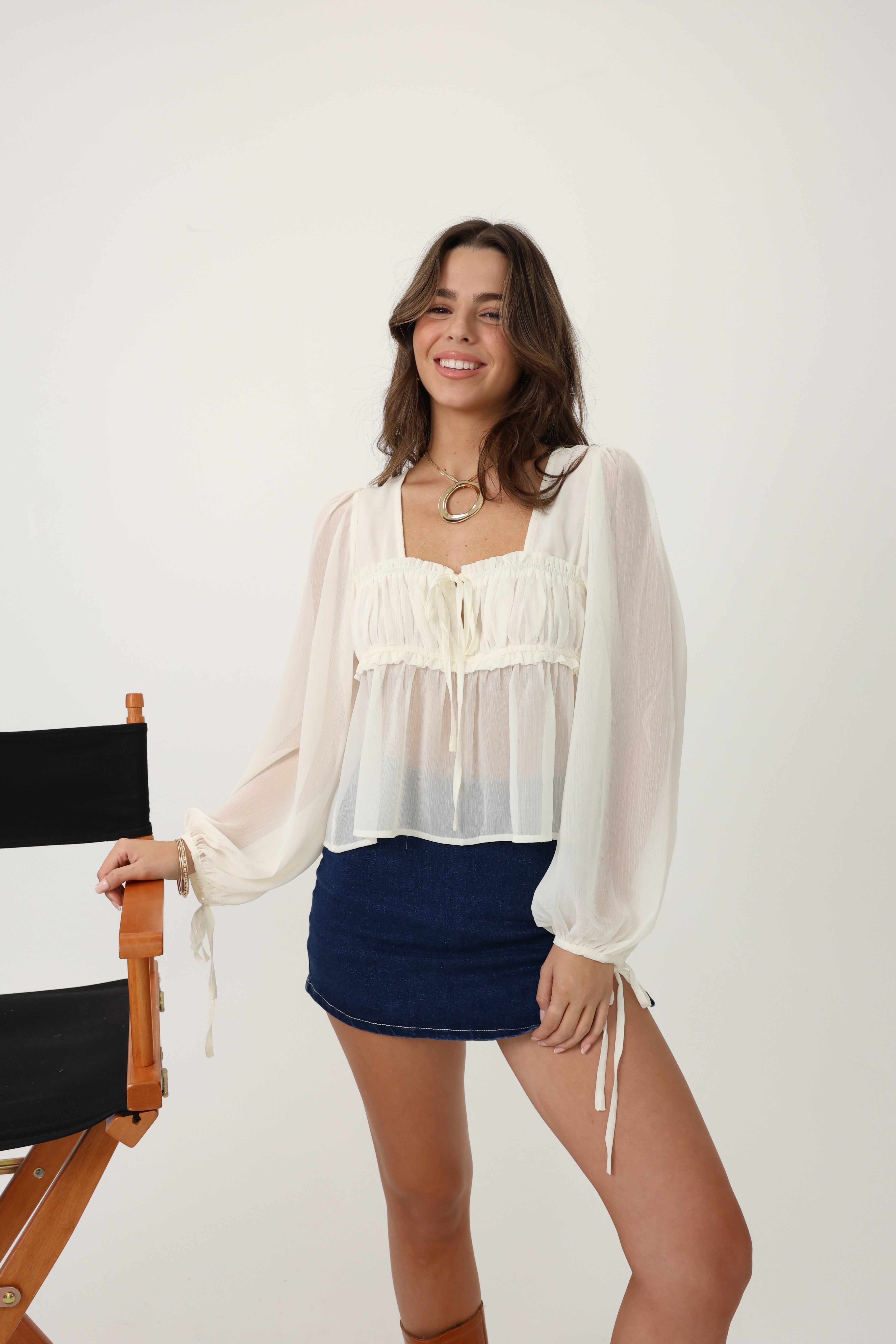 Woman wearing a white blouse and navy skirt standing next to a director's chair on a white background