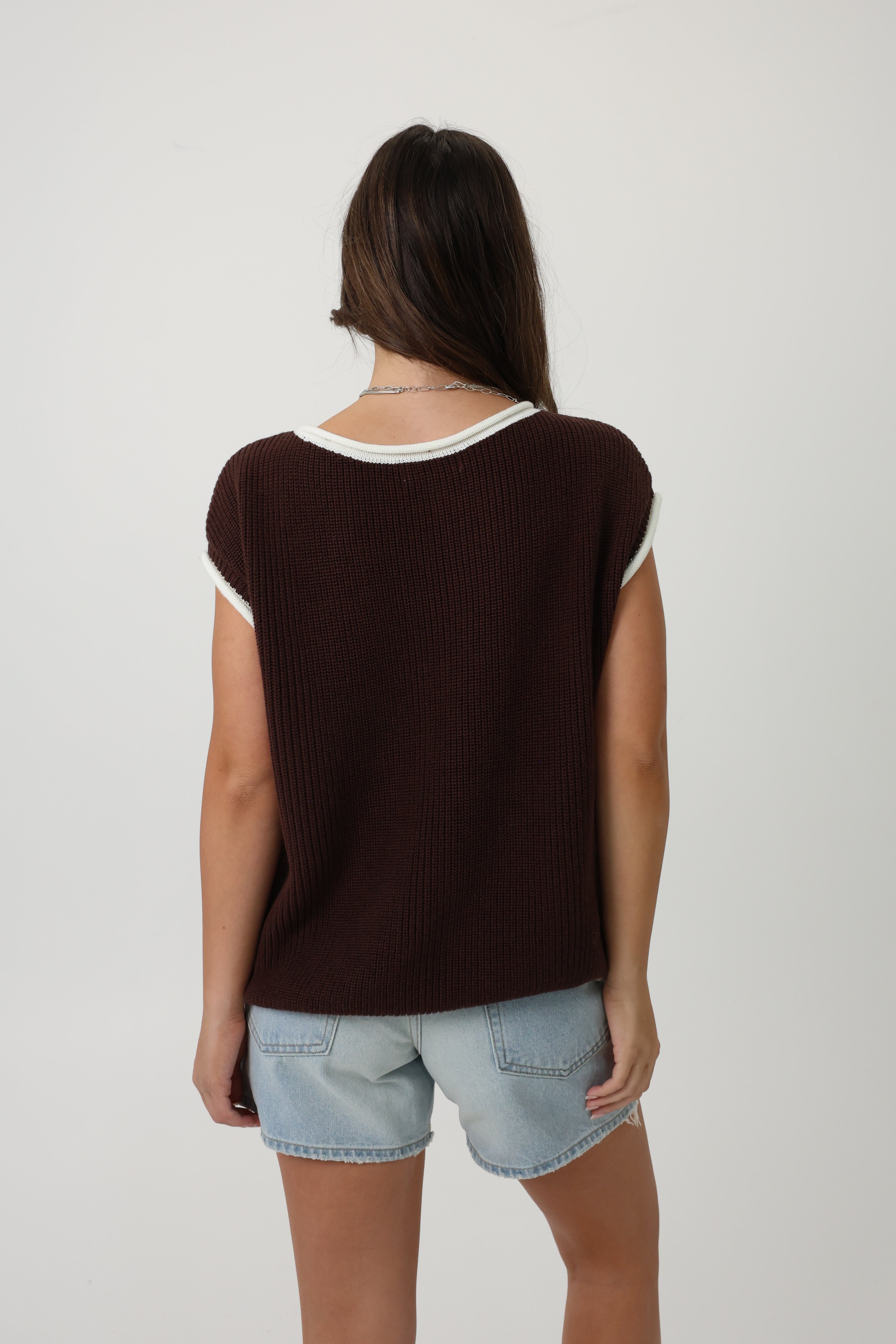 Person wearing a brown sleeveless top with white trim and light blue jeans shorts on a white background