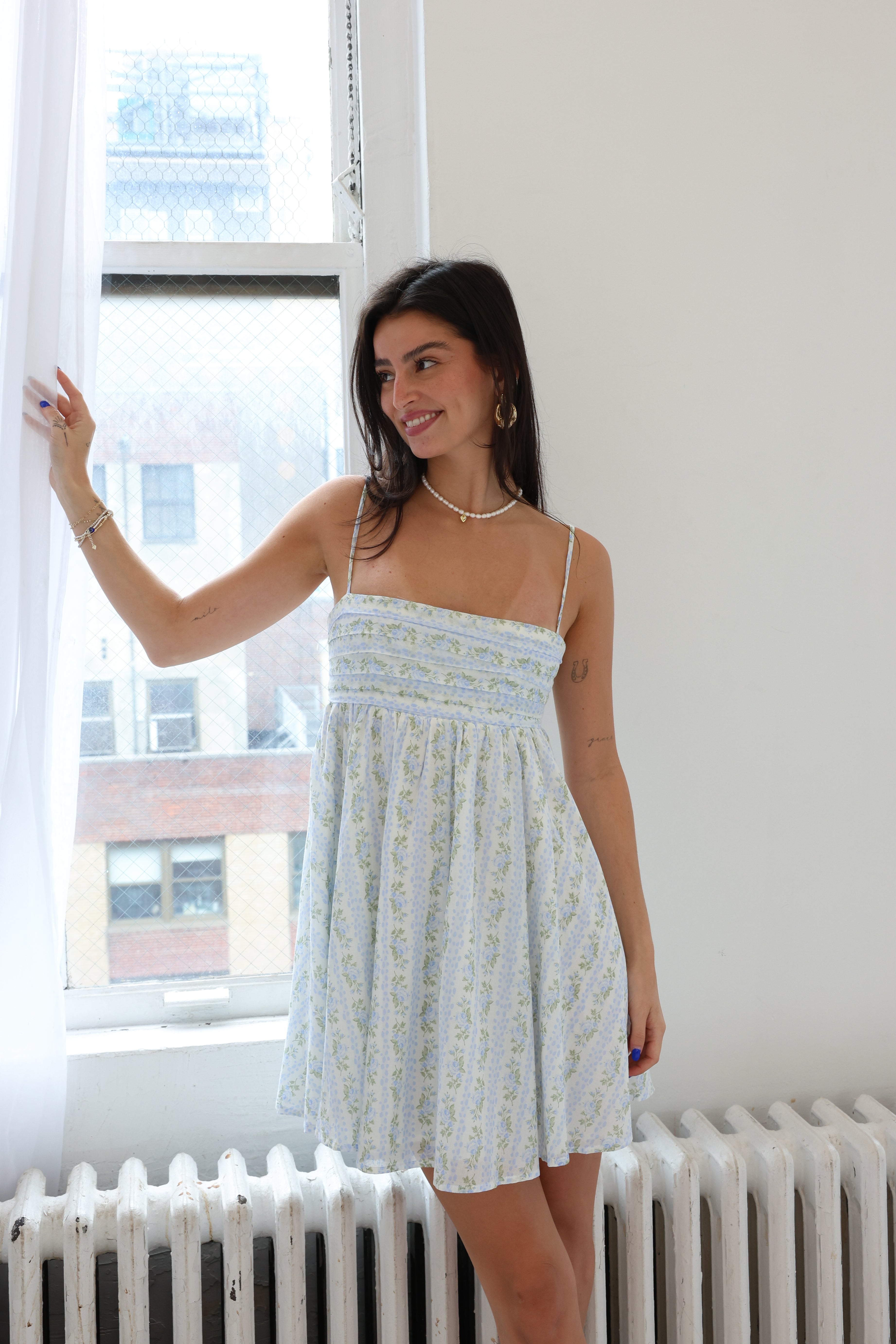 Woman in a white patterned dress standing by a window in a room with a radiator.