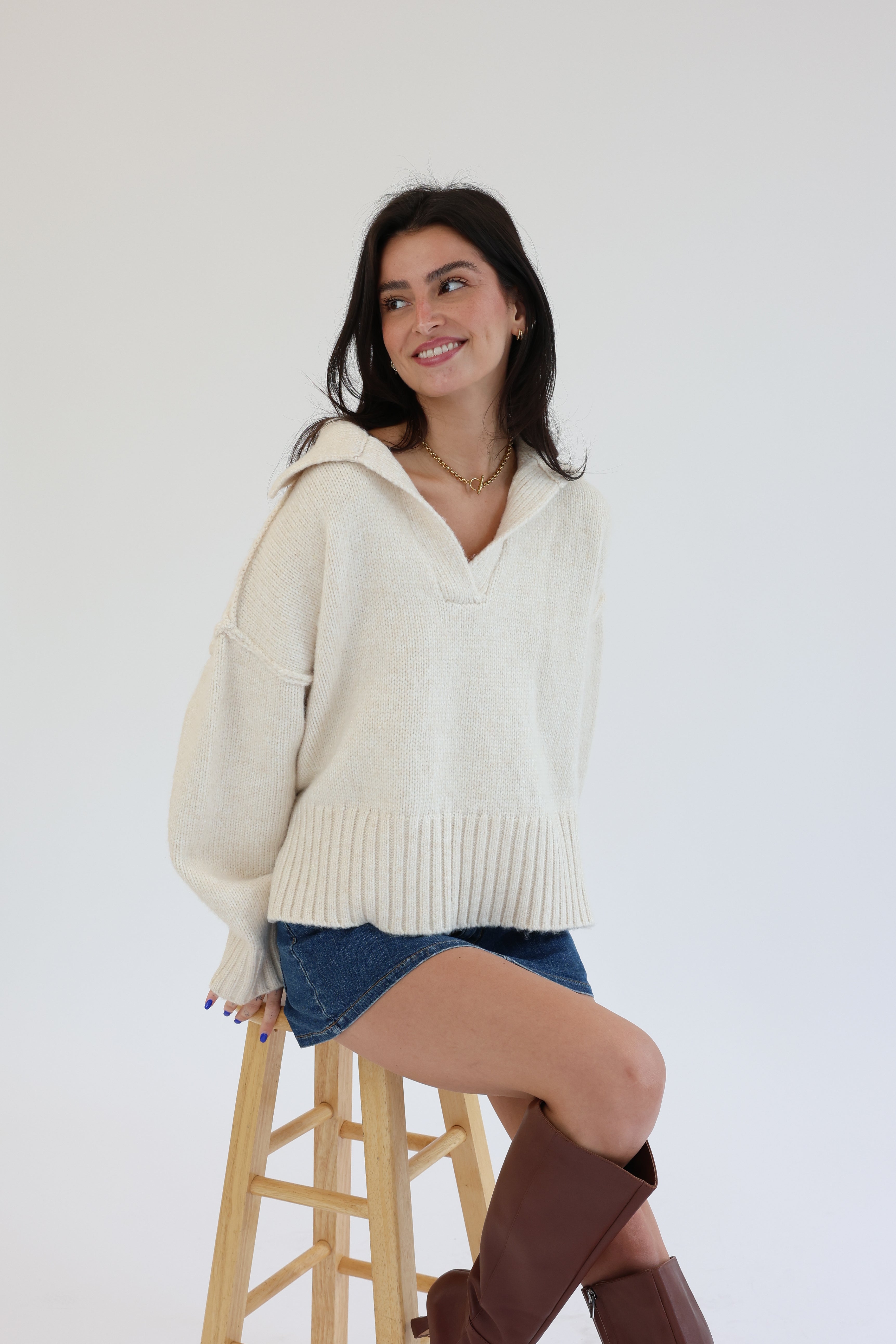 Woman wearing a cream sweater and denim shorts, sitting on a wooden stool against a white background