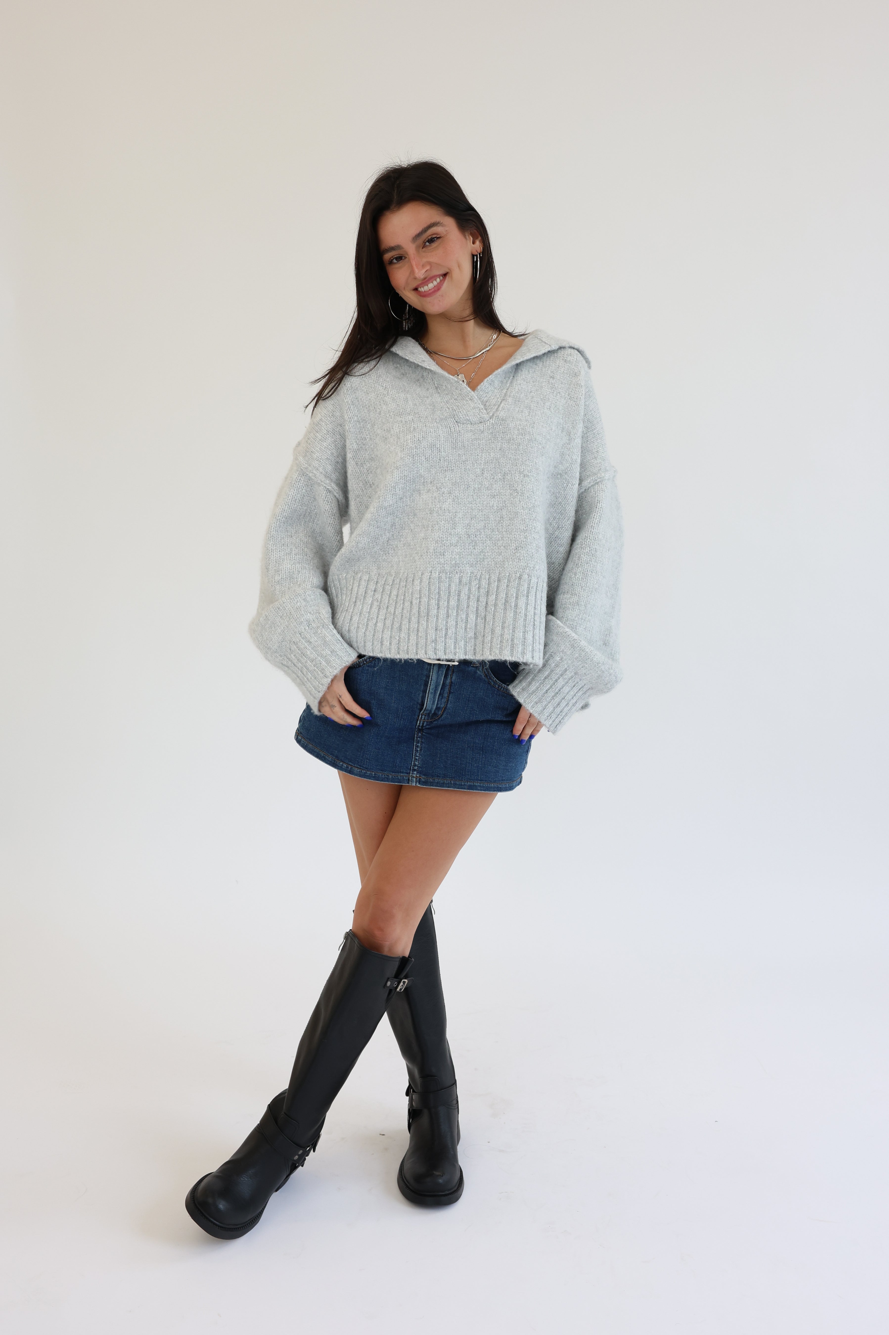 Woman wearing a light gray sweater, denim skirt, and black knee-high boots on a white background