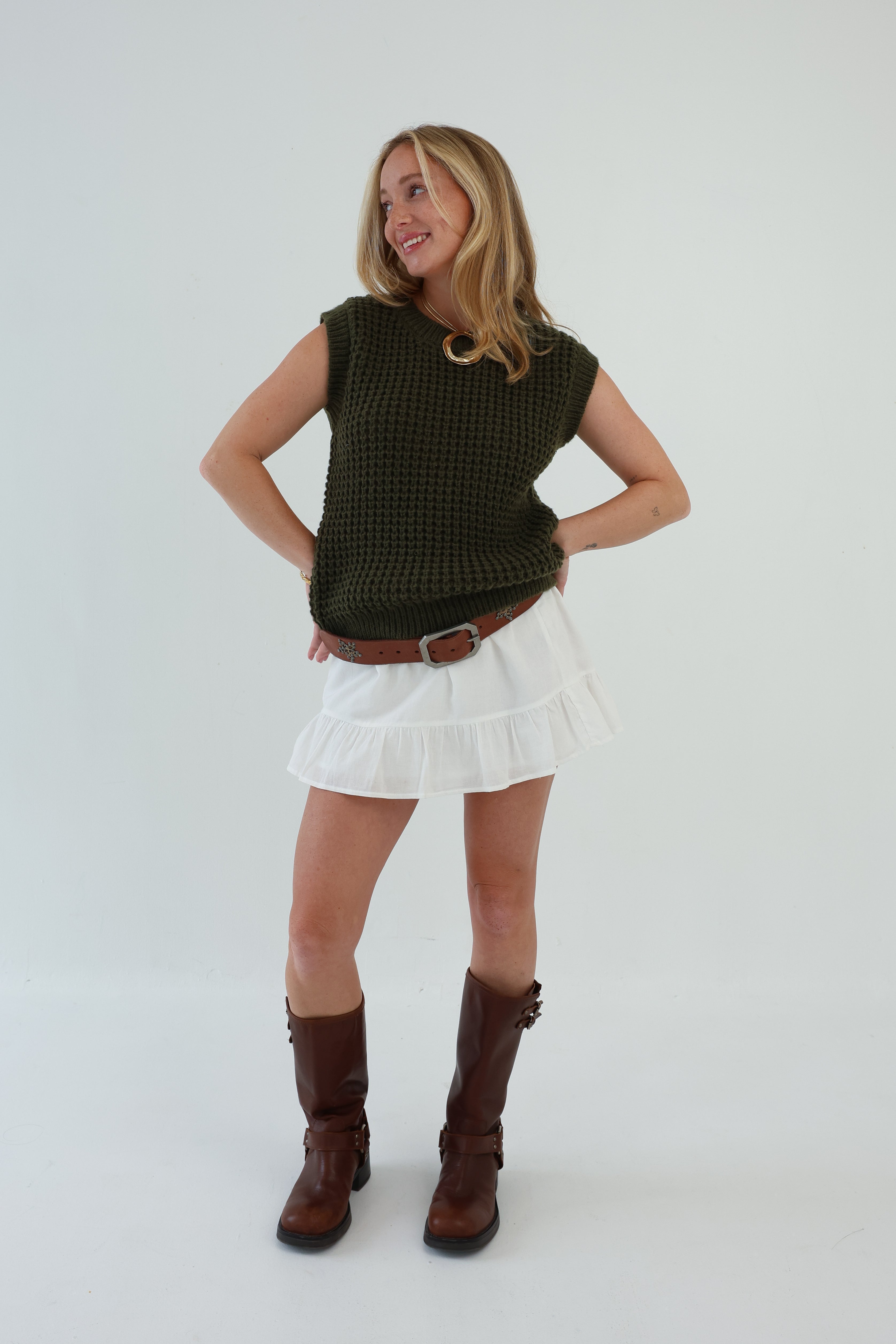 Woman wearing a green sleeveless top, white skirt, and brown boots on a white background