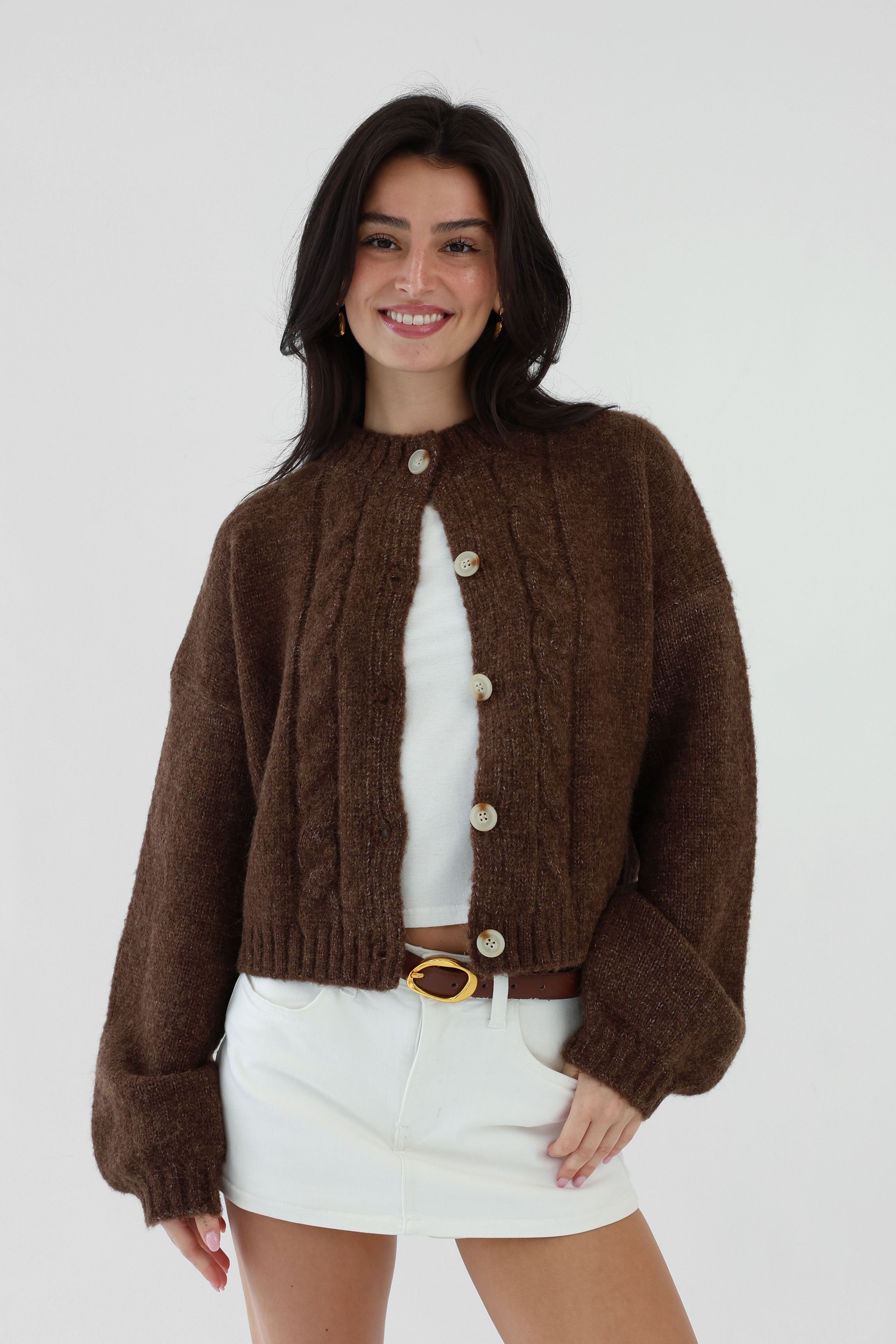 Woman wearing a brown knitted cardigan over a white top and skirt on a plain background