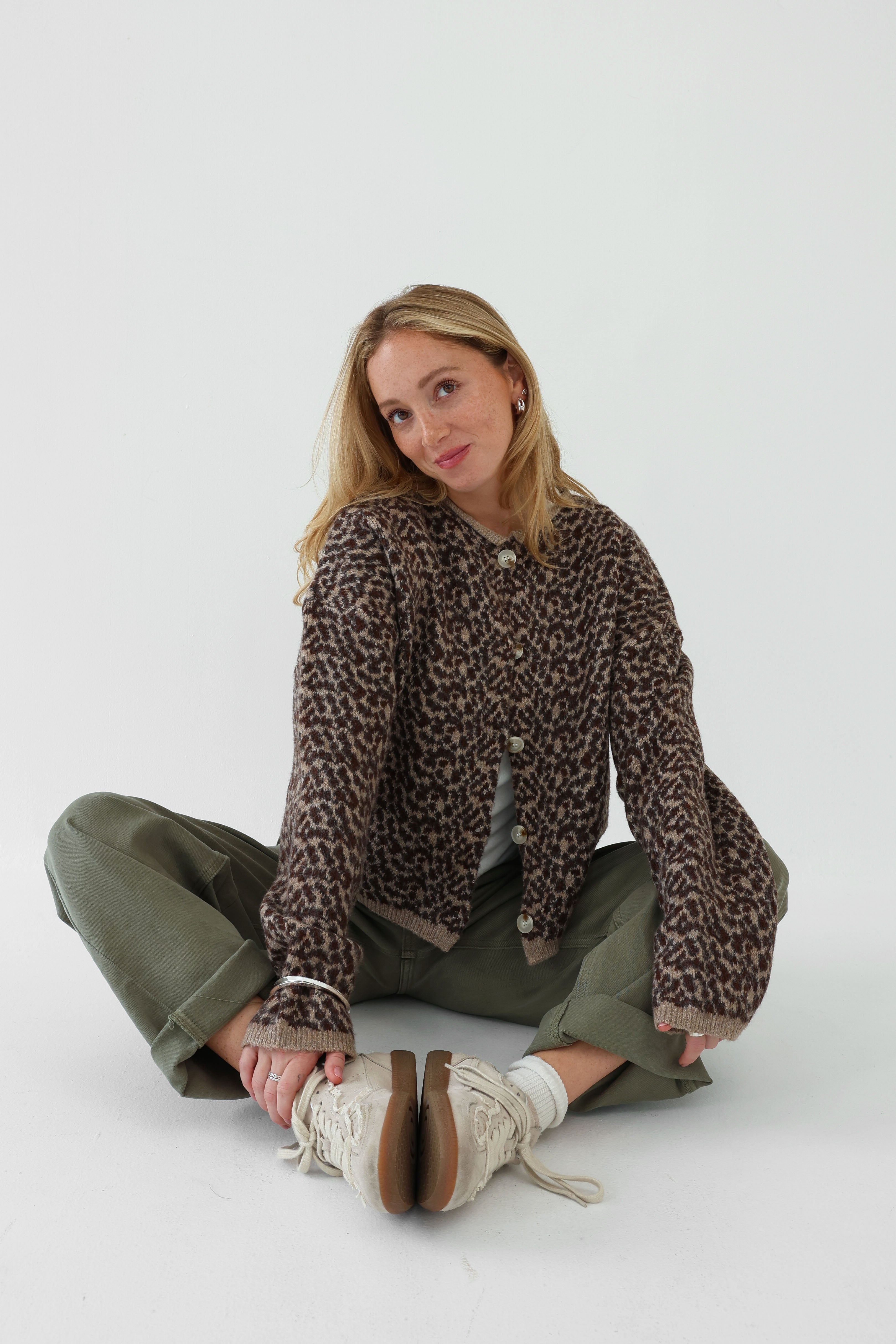 Woman wearing a leopard print sweater and green pants sitting on the floor against a white background
