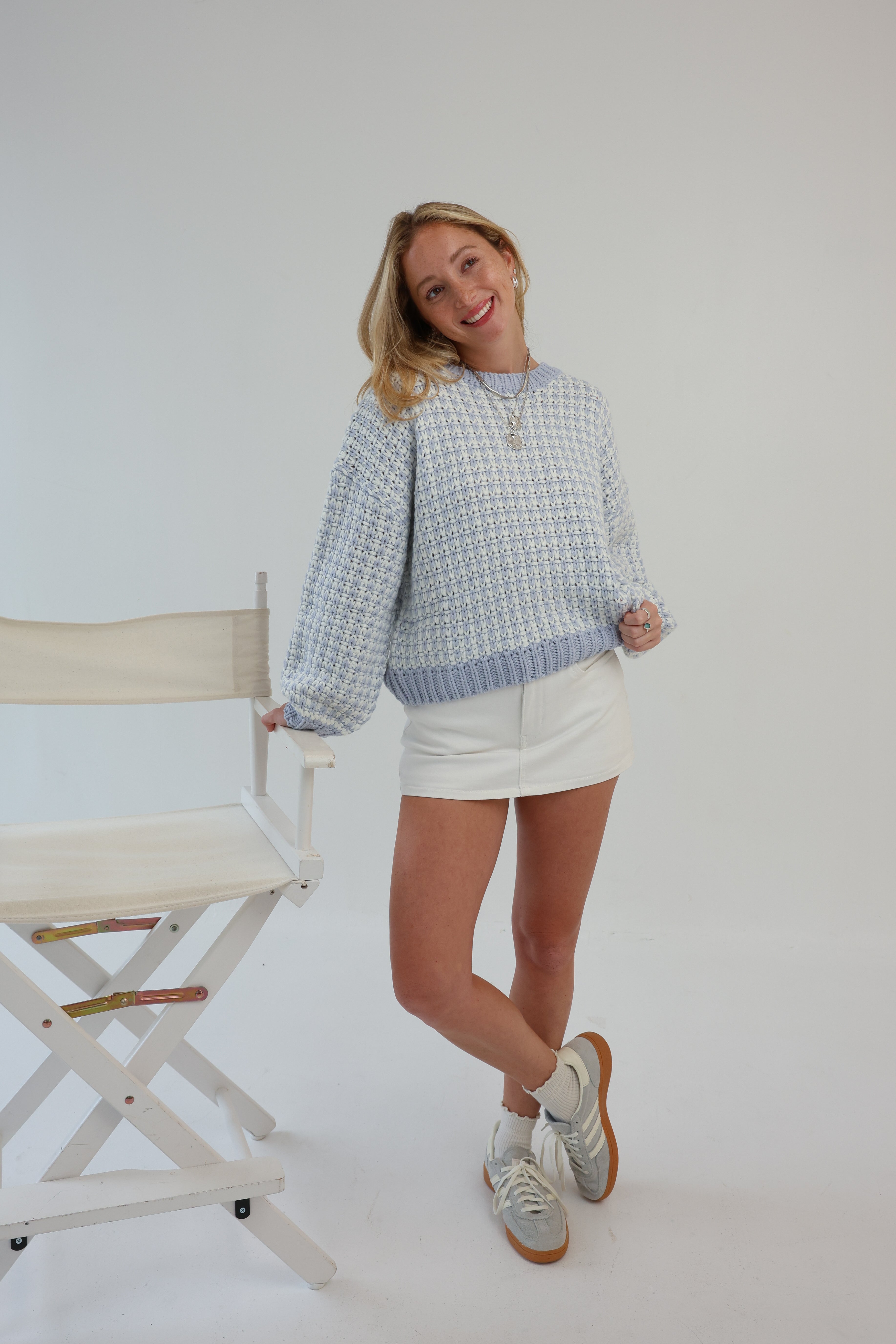 Woman wearing a patterned sweater and a white skirt standing next to a chair on a plain background