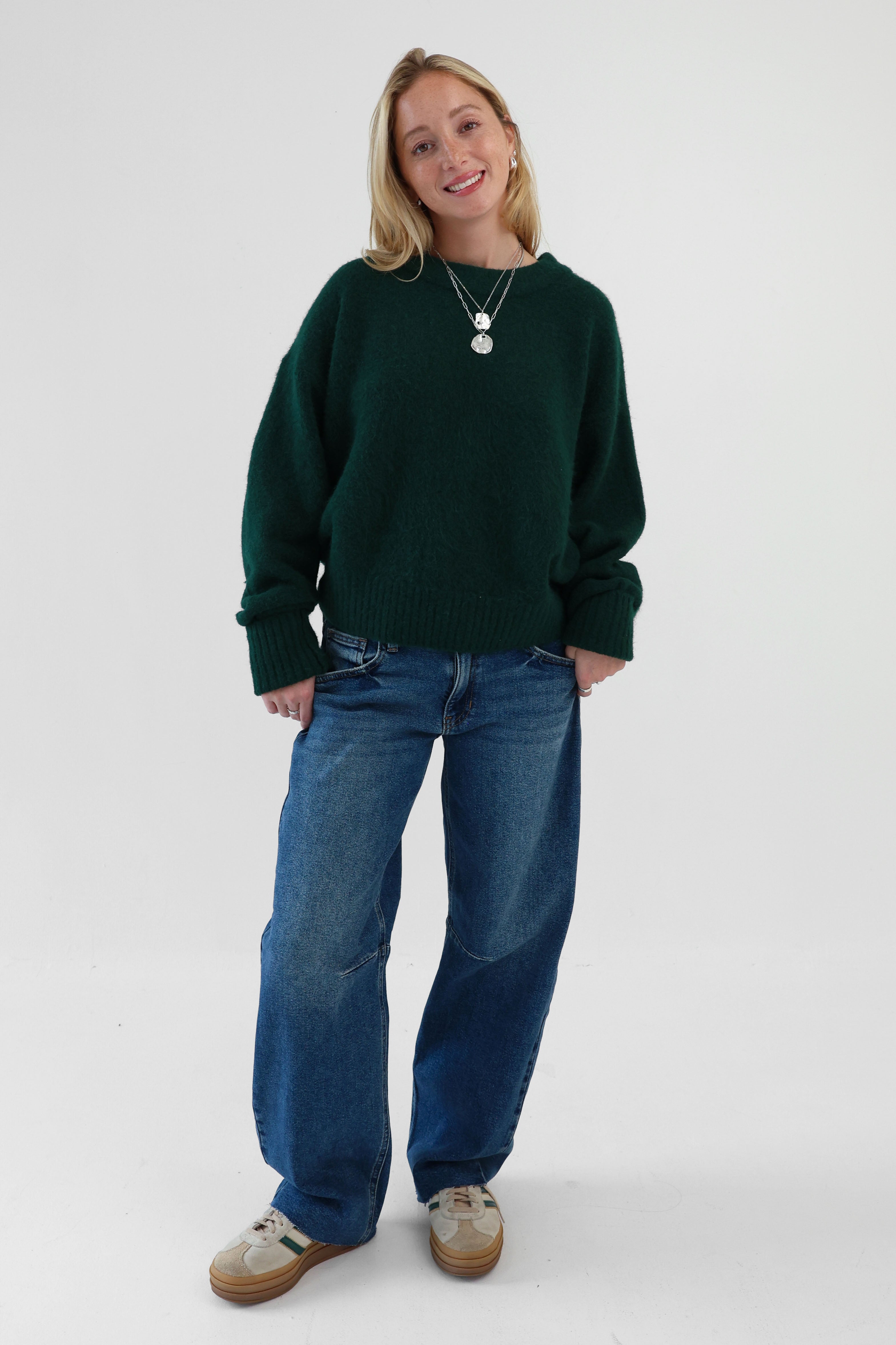 A woman wearing a green sweater and blue jeans against a white background